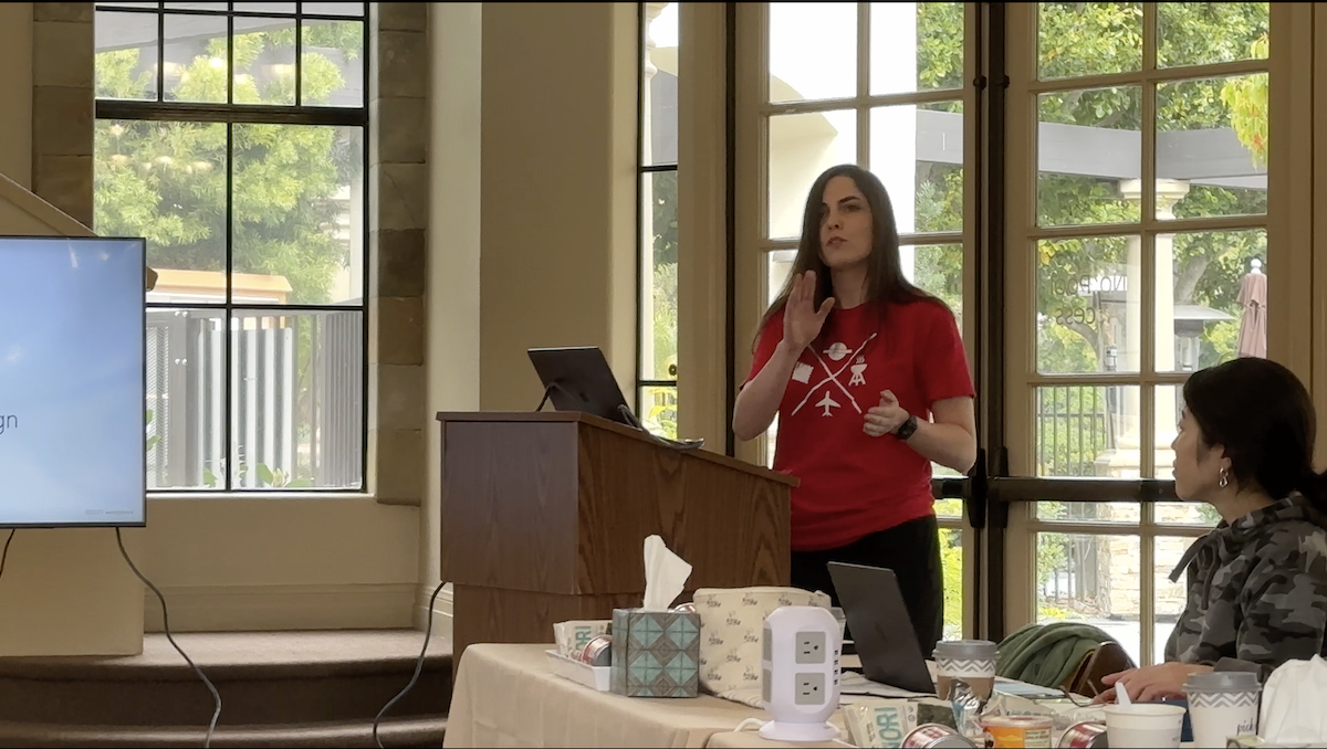 Renata at a lectern giving a design talk, with the audience seated nearby.