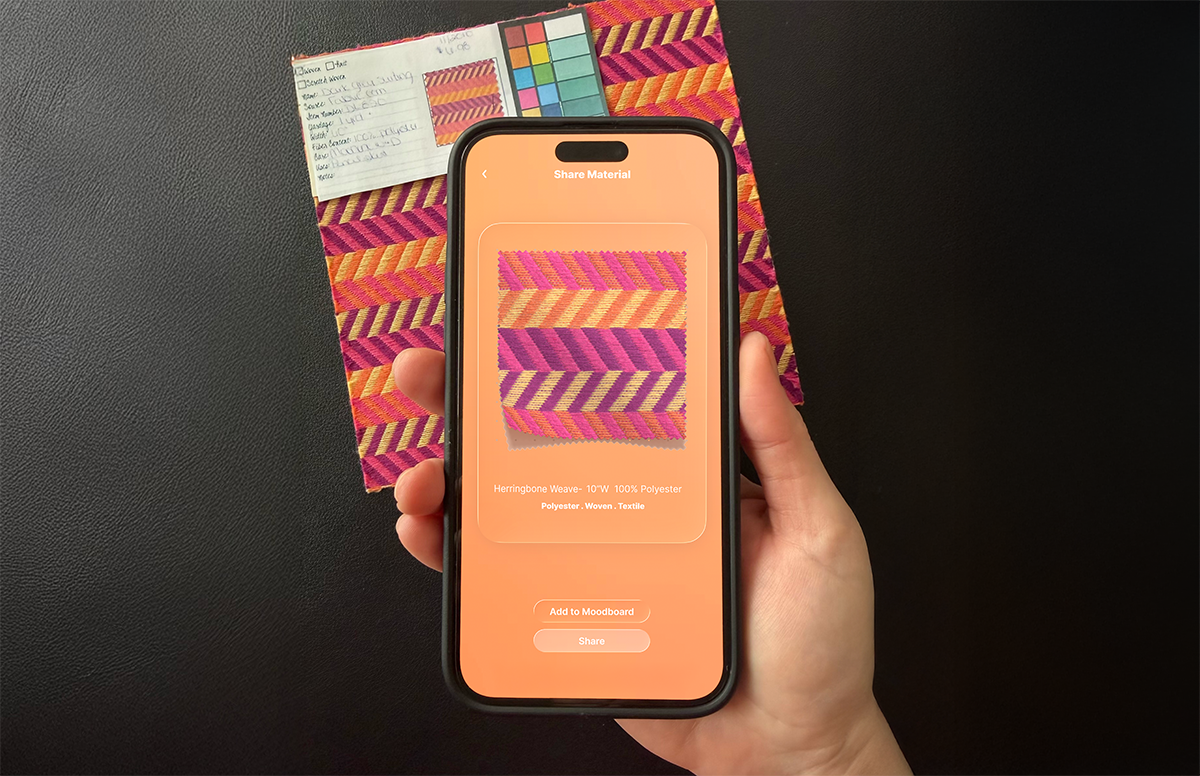 A hand holding a phone that is capturing a patterned woven fabric, with the physical swatch card behind it.