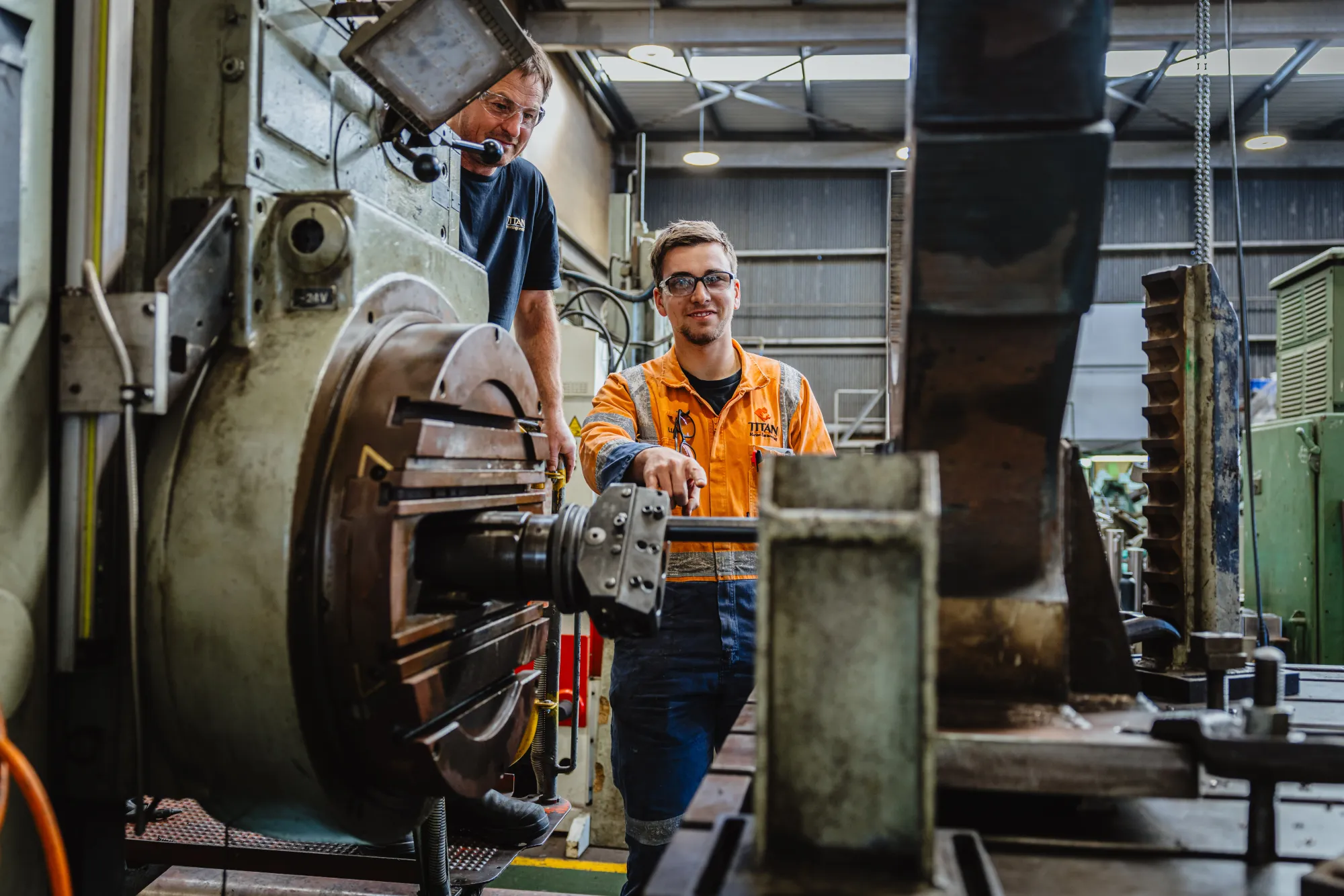 A coach watches an apprentice machine a metal component