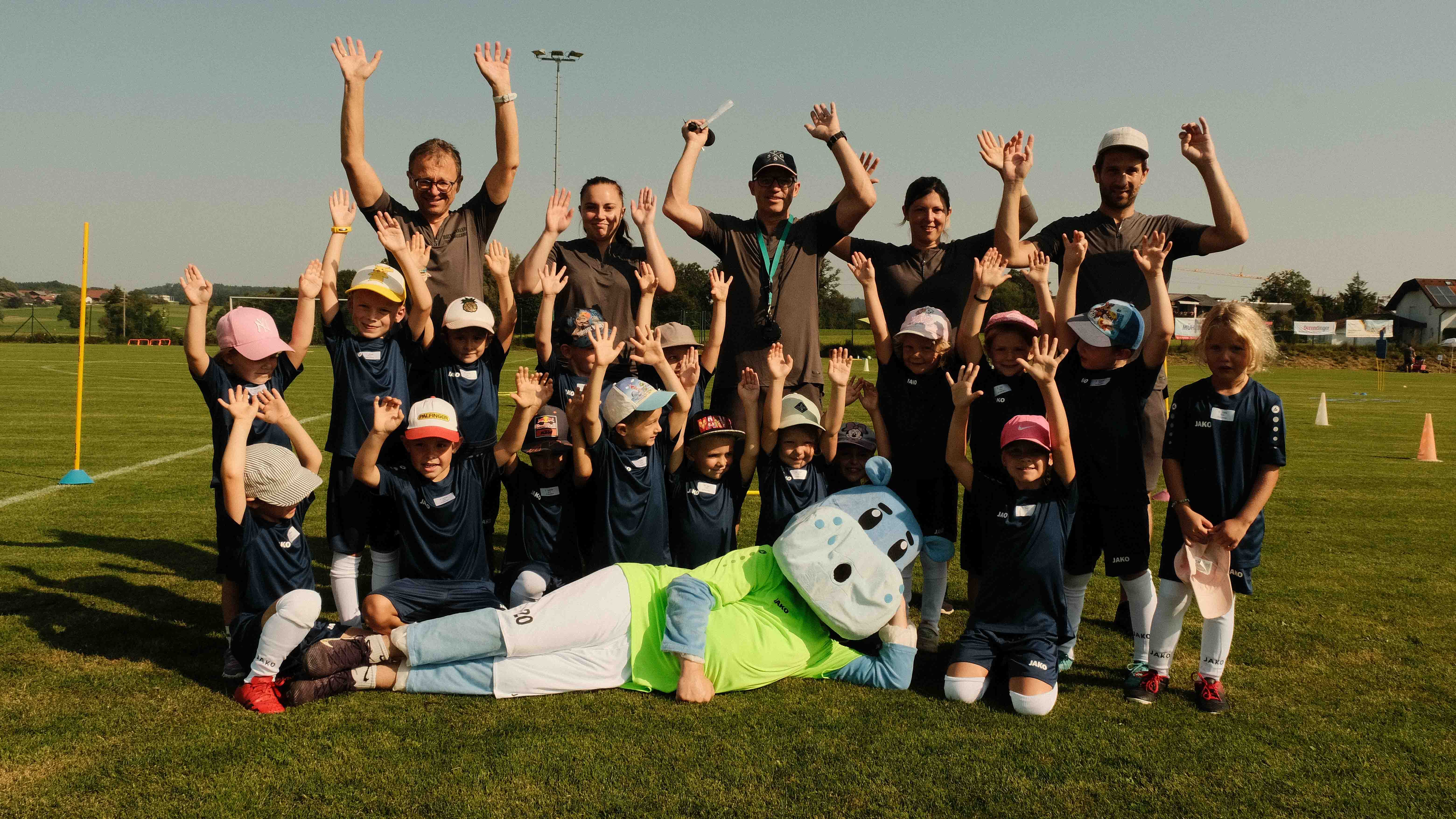 Kinder und Betreuer in Sportkleidung posieren lachend auf einem Fußballfeld, mit einer liegenden Nilpferd-Maskottchenfigur vorne.
