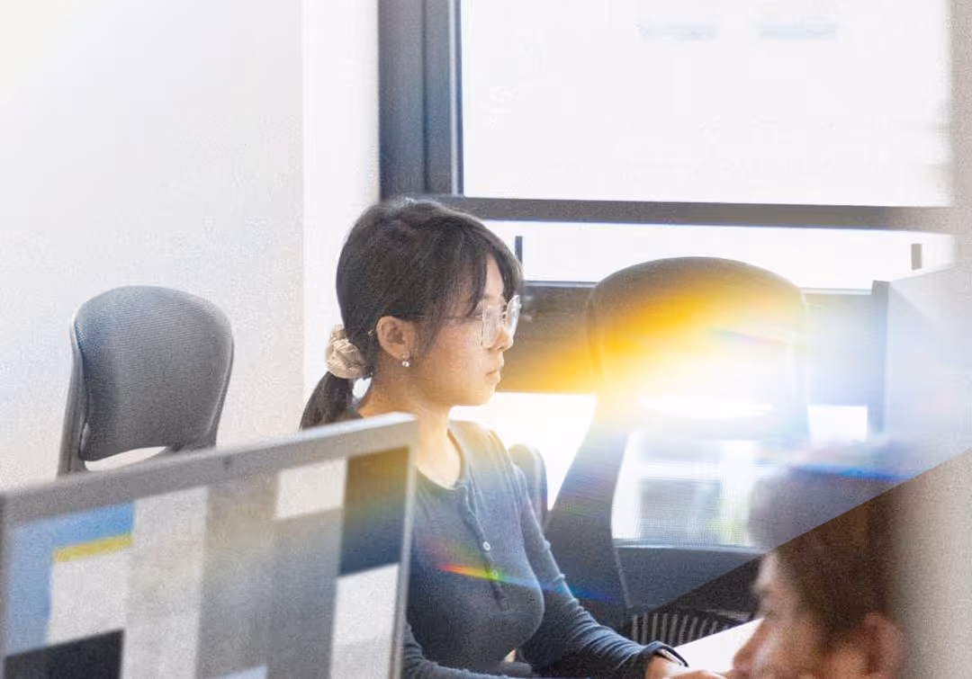 Woman with glasses working on a computer in a bright office with black chairs and window behind her.