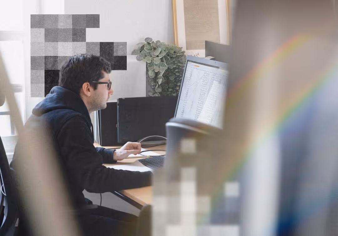 Man with glasses working on a computer at a desk with plants and framed art in the background.