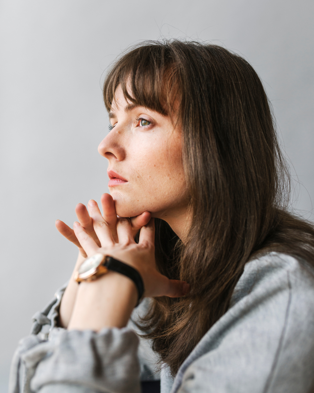 Side profile of a woman with brown hair resting her chin on her interlaced fingers, looking thoughtful.