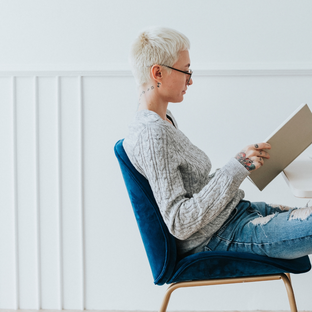 Person with short blonde hair and glasses reading a book while sitting on a blue chair.