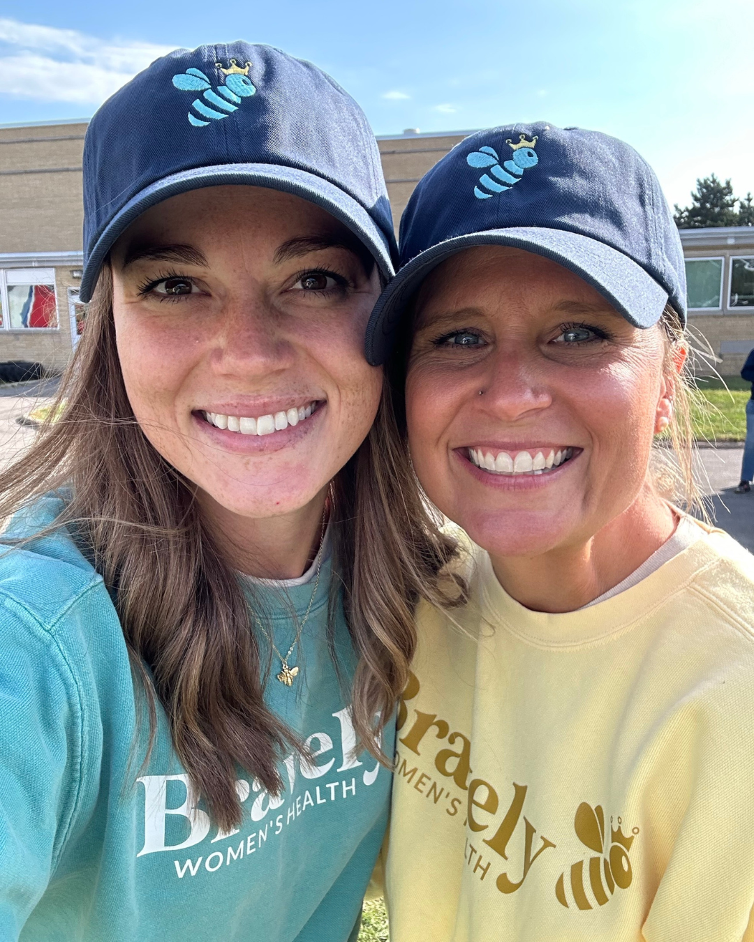 Two smiling women outdoors wearing matching navy caps with blue bee logos and branded sweatshirts.