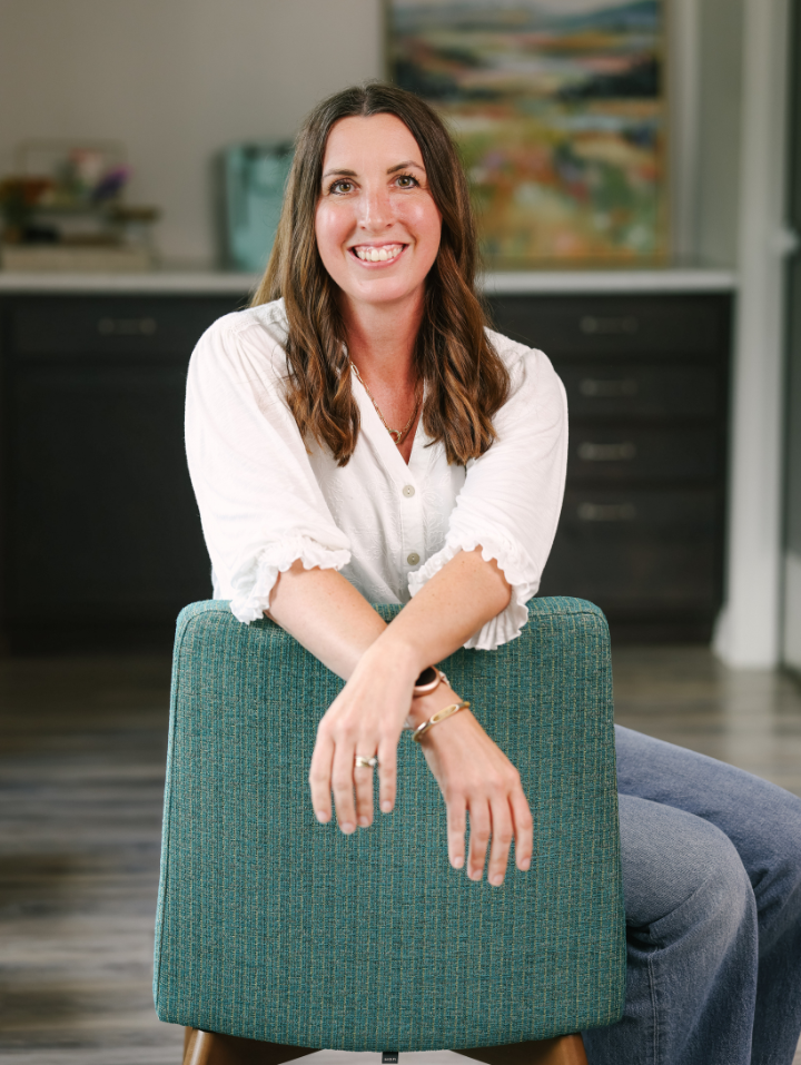 Smiling woman with brown hair sits backward on teal chair in a modern room with wooden floor.