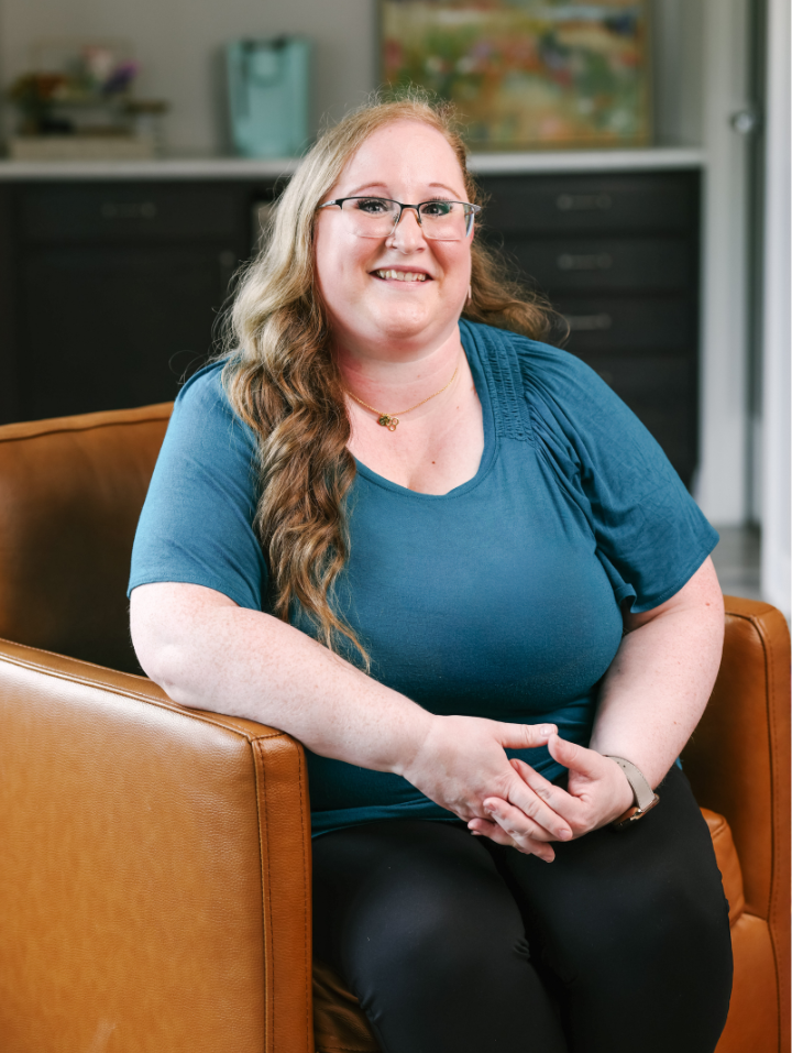 Woman with long curly hair and glasses, smiling while sitting on a brown leather chair indoors.