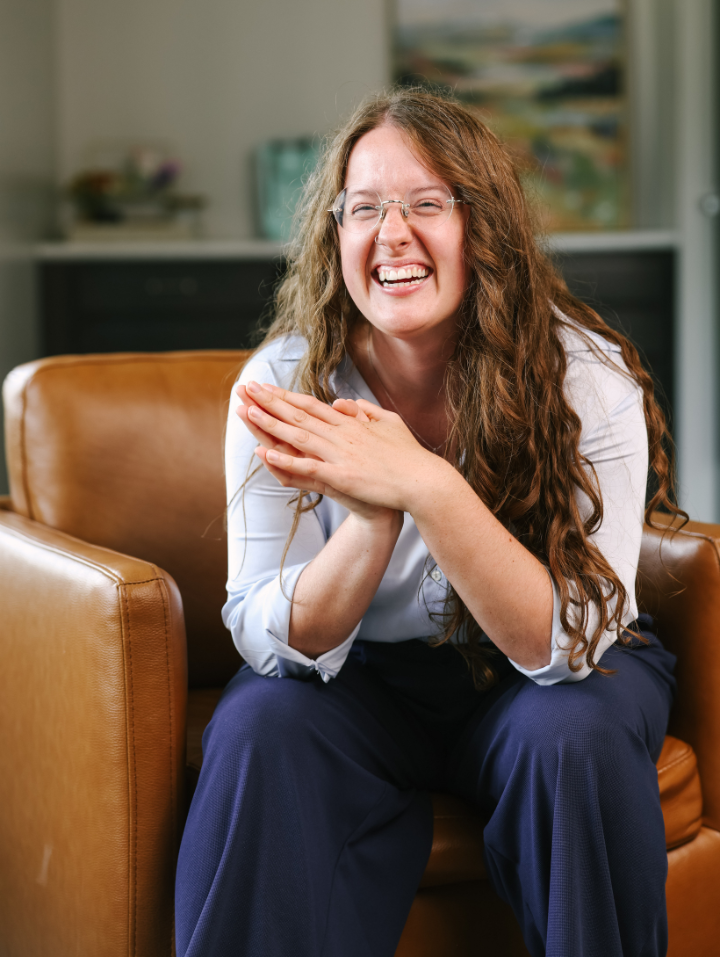 Smiling woman with long curly hair and glasses sitting on a brown leather chair, hands clasped.
