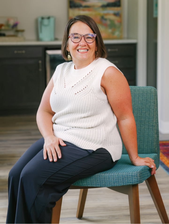 Smiling woman with short brown hair and glasses sitting on a teal chair in a home setting.