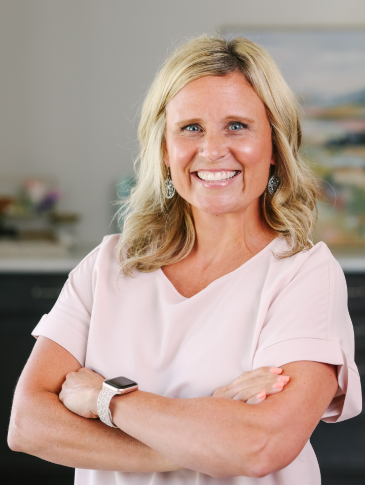 Smiling woman with blonde hair wearing a light pink blouse and a smartwatch, arms crossed.