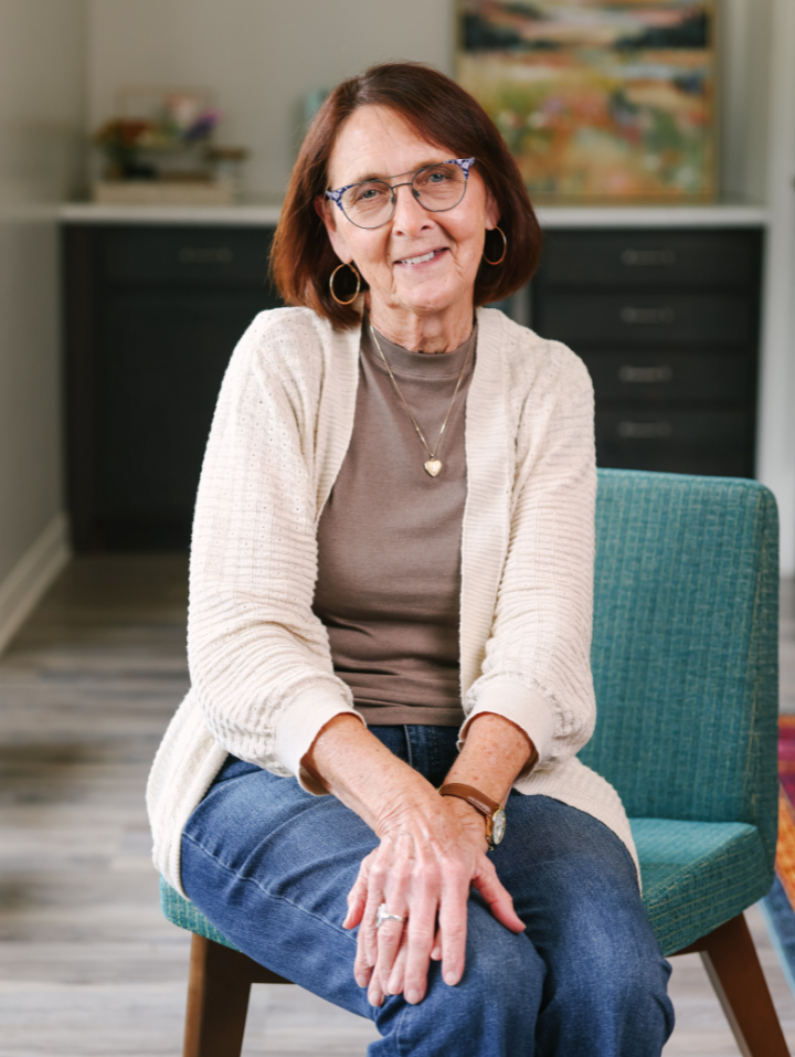 Older woman with glasses, beige sweater, and blue jeans sitting on teal chair smiling gently.