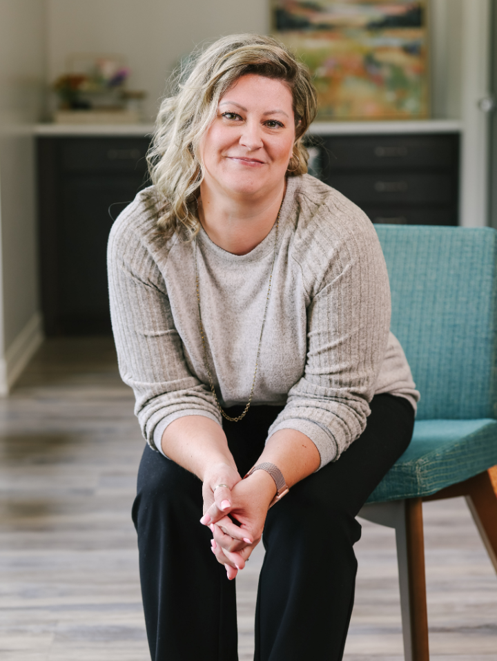 Smiling woman with wavy hair seated, wearing a beige sweater and black pants in a modern room.