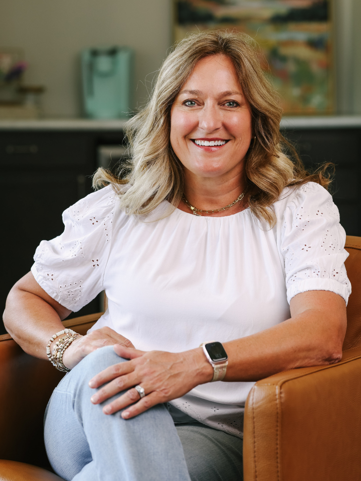 Smiling woman with wavy blond hair sitting in a brown chair wearing a white blouse and light jeans.