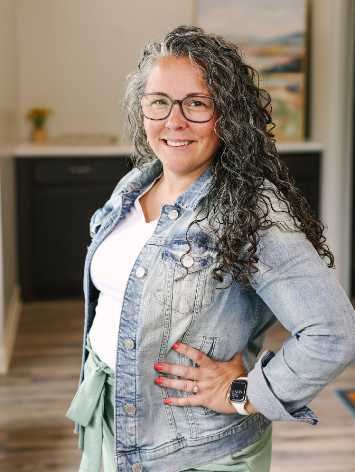 Smiling woman with curly gray hair wearing glasses, denim jacket, and smartwatch posing indoors.