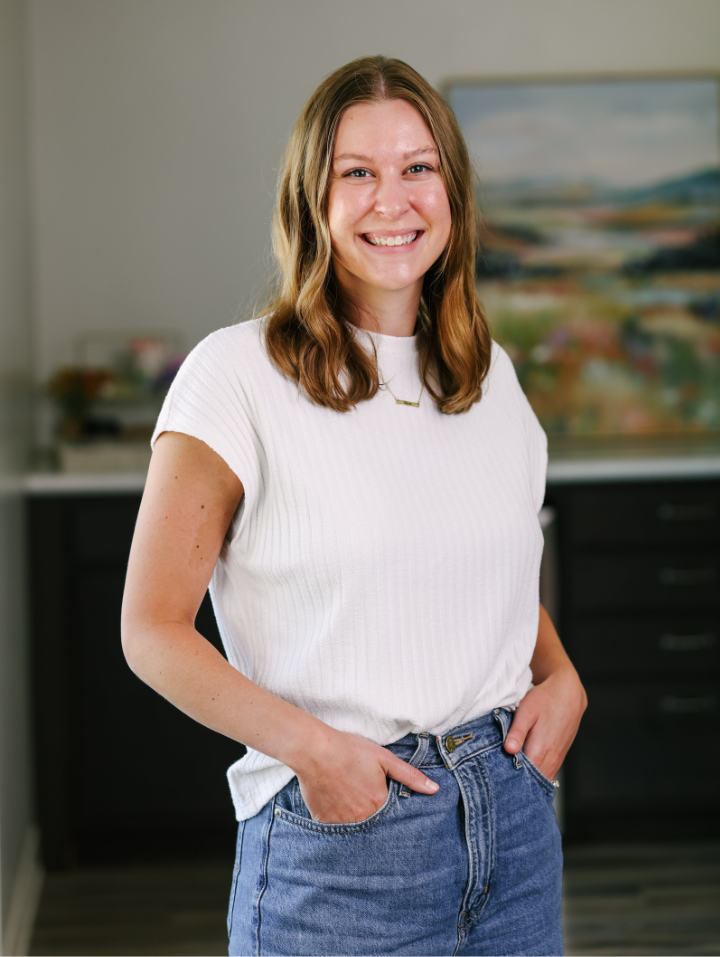 Smiling woman with wavy light brown hair wearing a white shirt and blue jeans with hands in pockets.