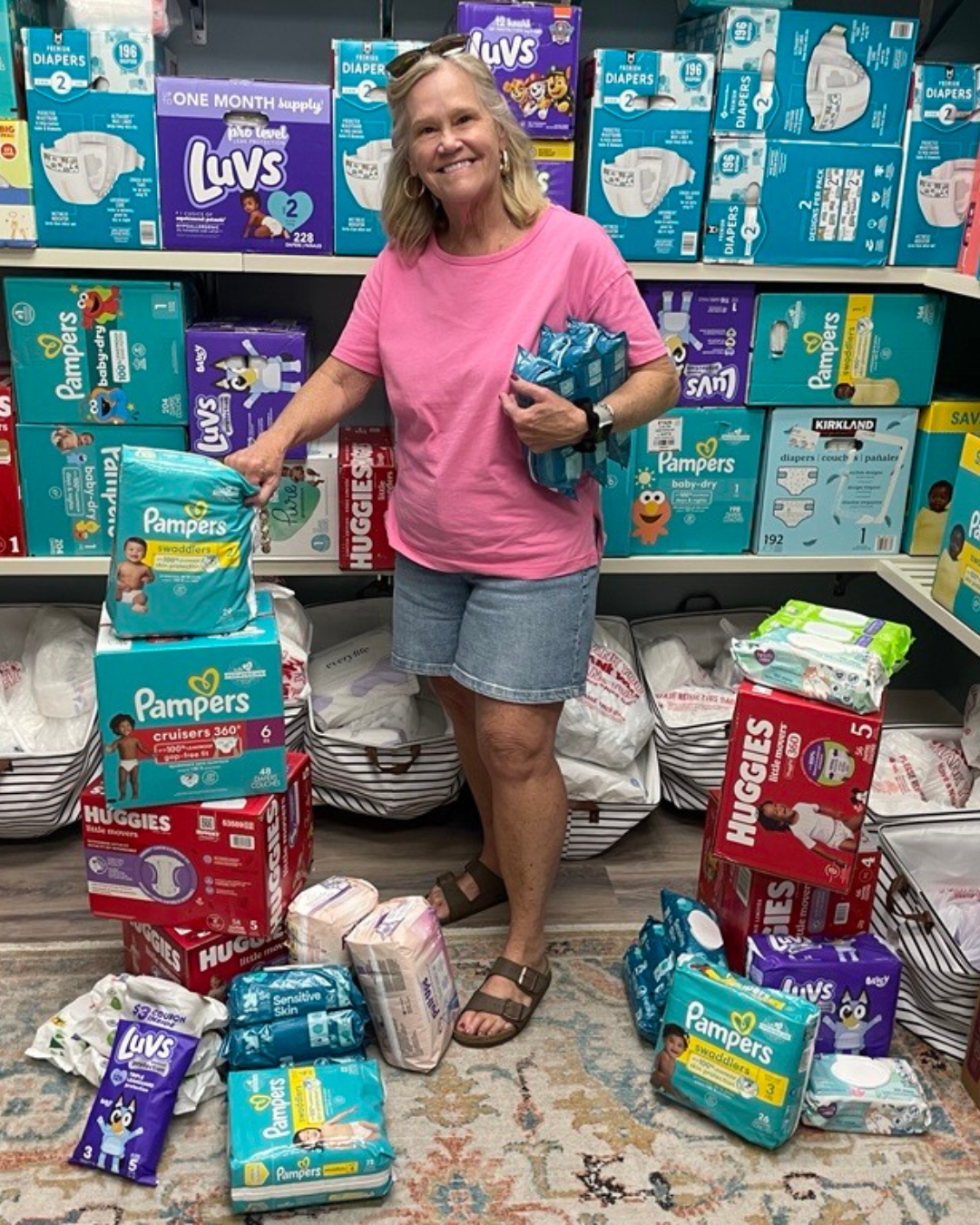 Smiling woman in pink shirt and denim shorts among various diaper packages in a room.