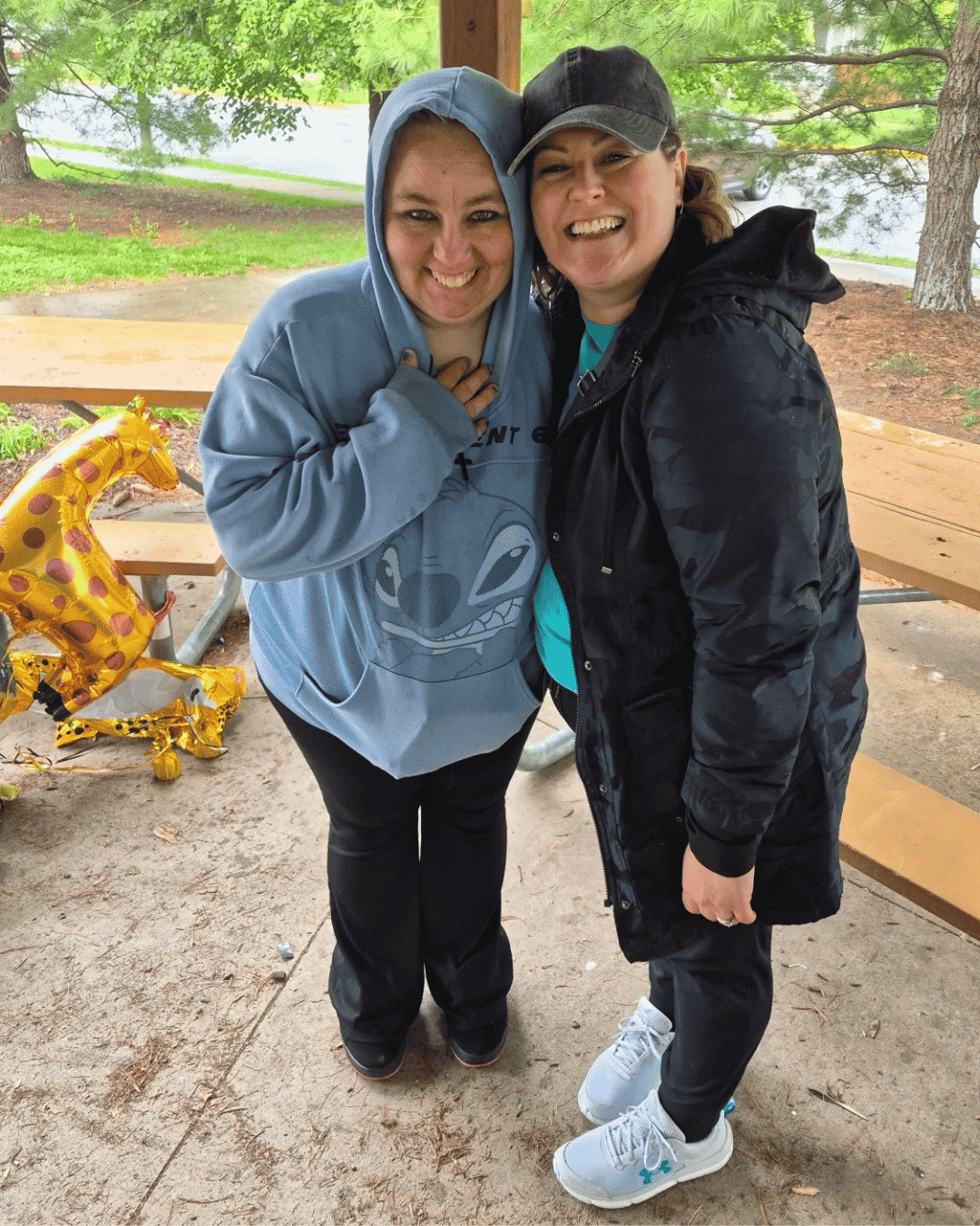 Two smiling women outdoors wearing matching navy caps with blue bee logos and branded sweatshirts.