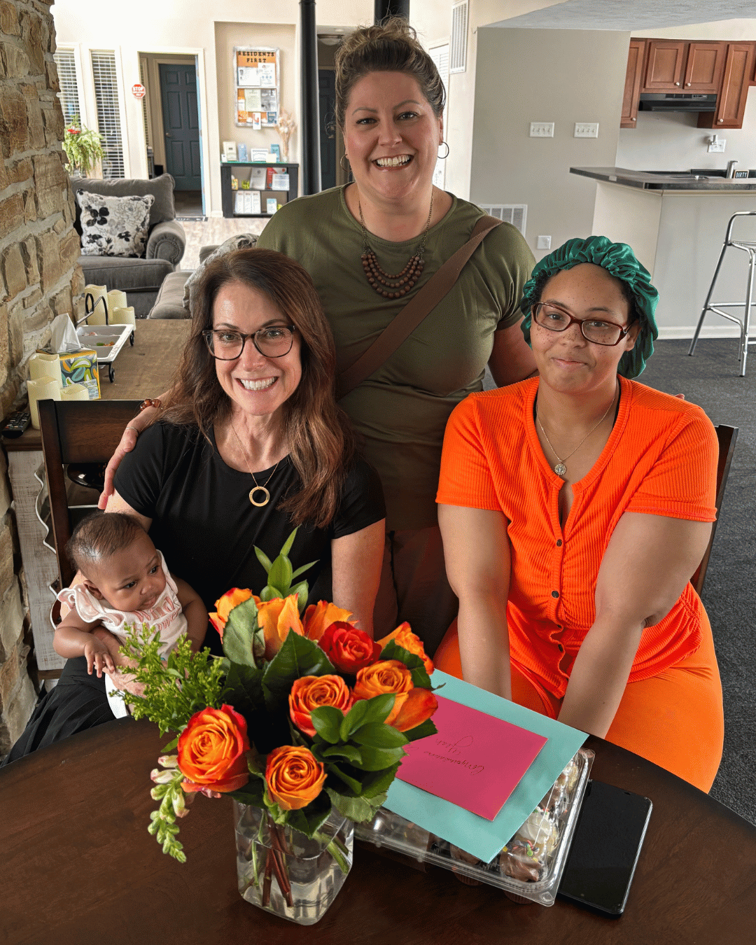 Three smiling women and a baby sitting at a table with orange roses and greeting cards.