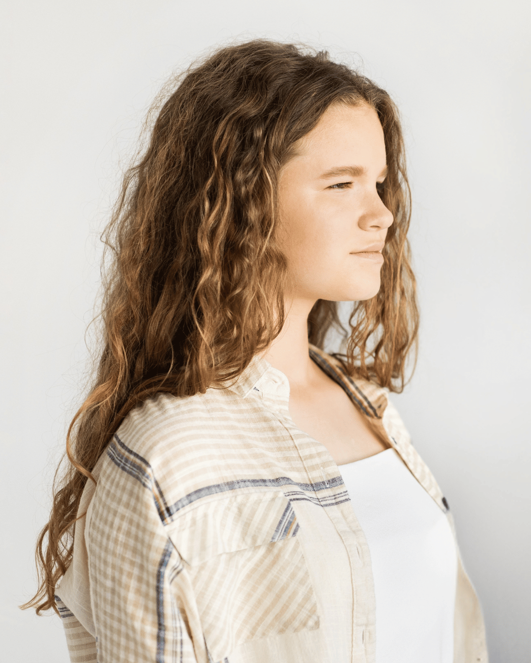 Profile of a young woman with long curly hair wearing a light plaid shirt and white top.