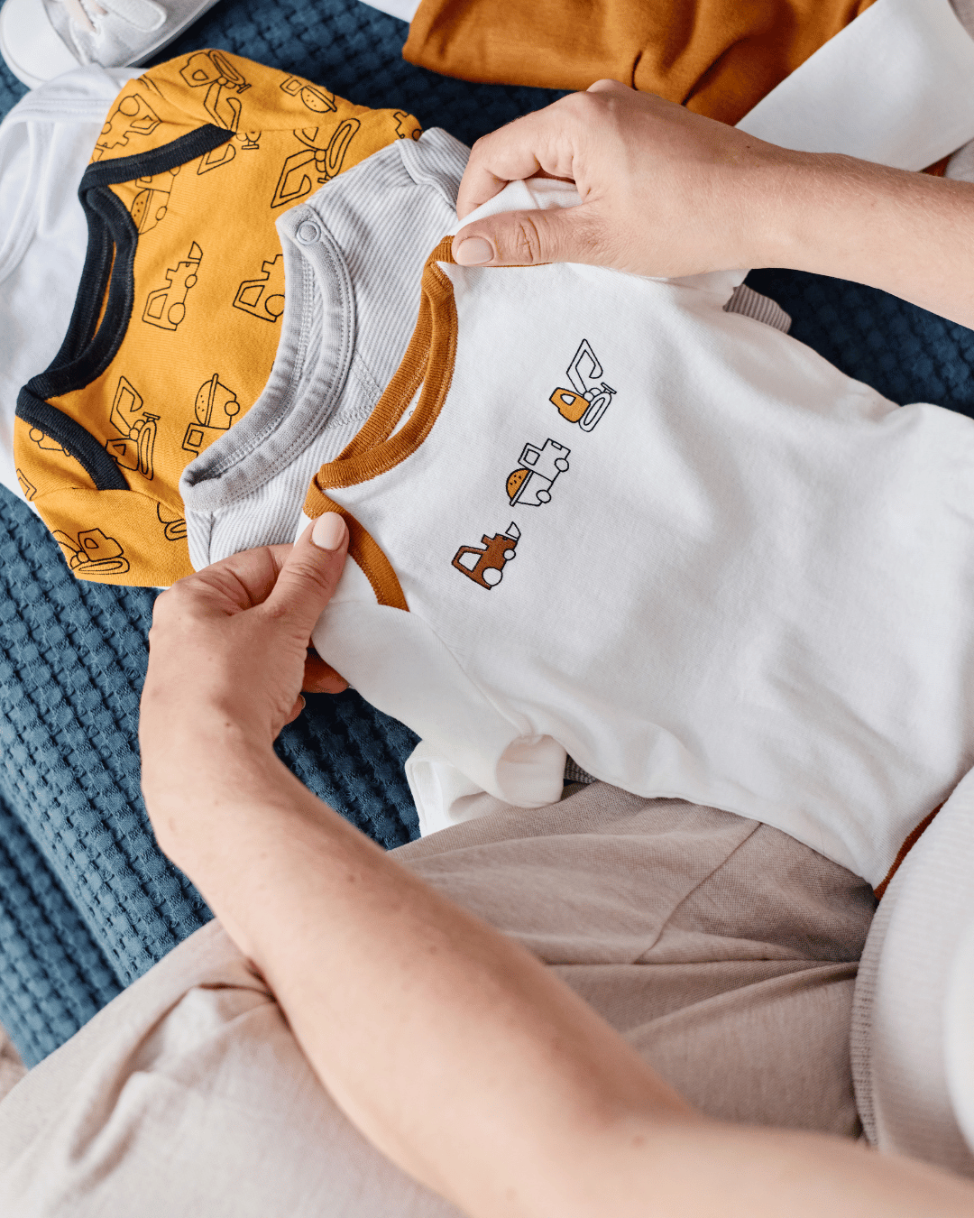A pregnant woman sitting on a bed, looking at baby shoes and sitting next to a pile of baby clothes.