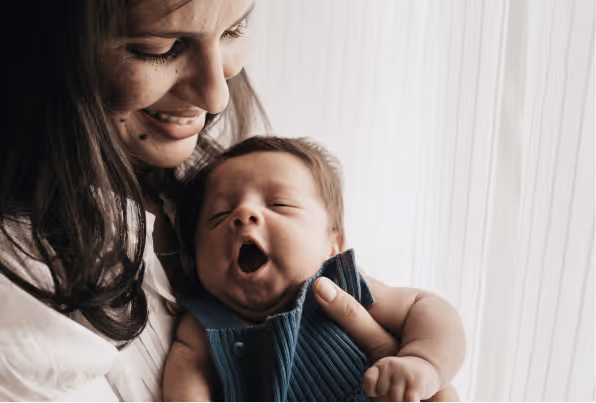 A woman holding her baby while smiling, sitting next to a window.