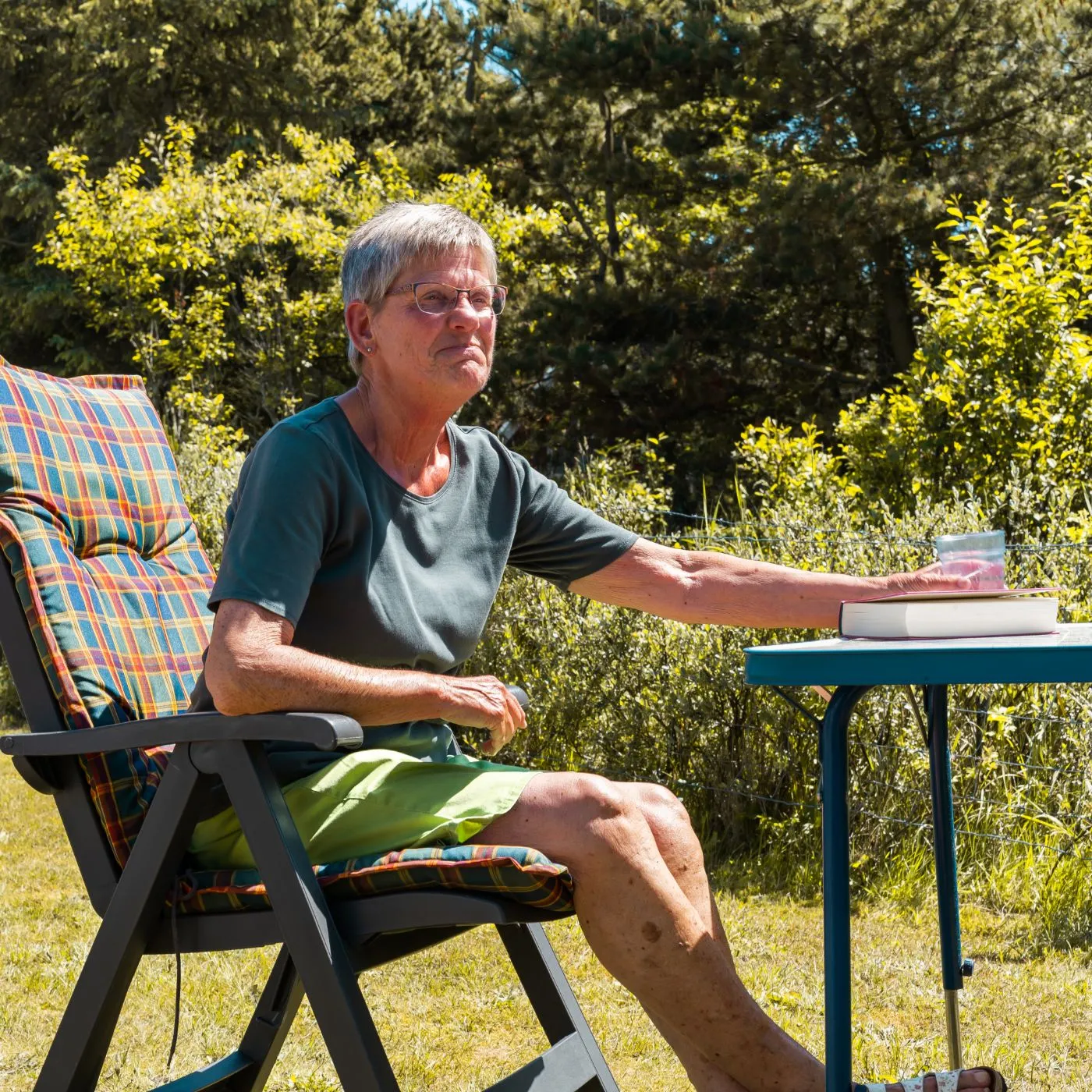 Ældre kvinde sidder udendørs på en stol med ternet pude, rækker ud efter en plastik kop på et bord med en bog, omgivet af grønne buske og træer.