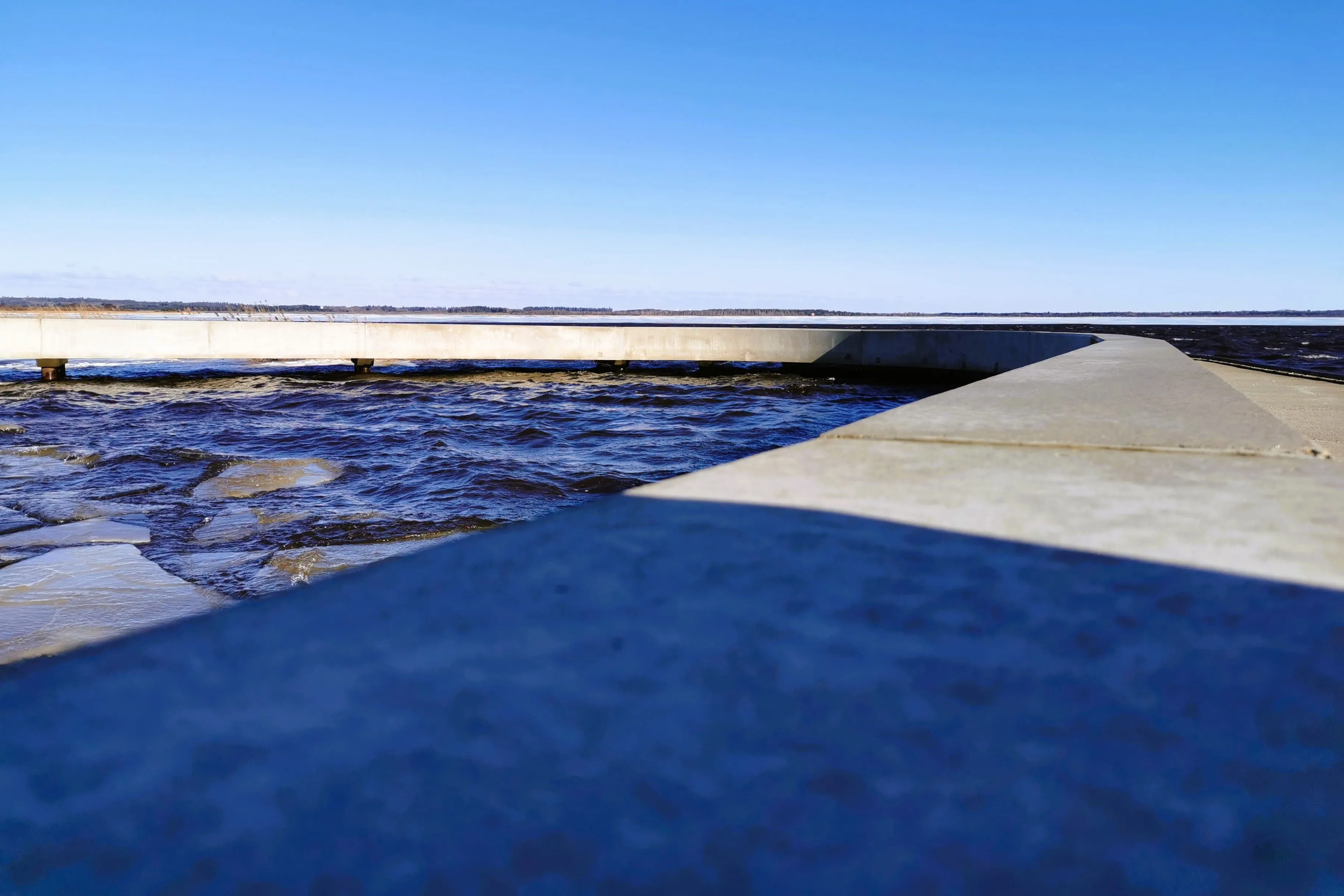 Betonstaumauer in einem See mit blauem Himmel und ferner Landschaft am Horizont.