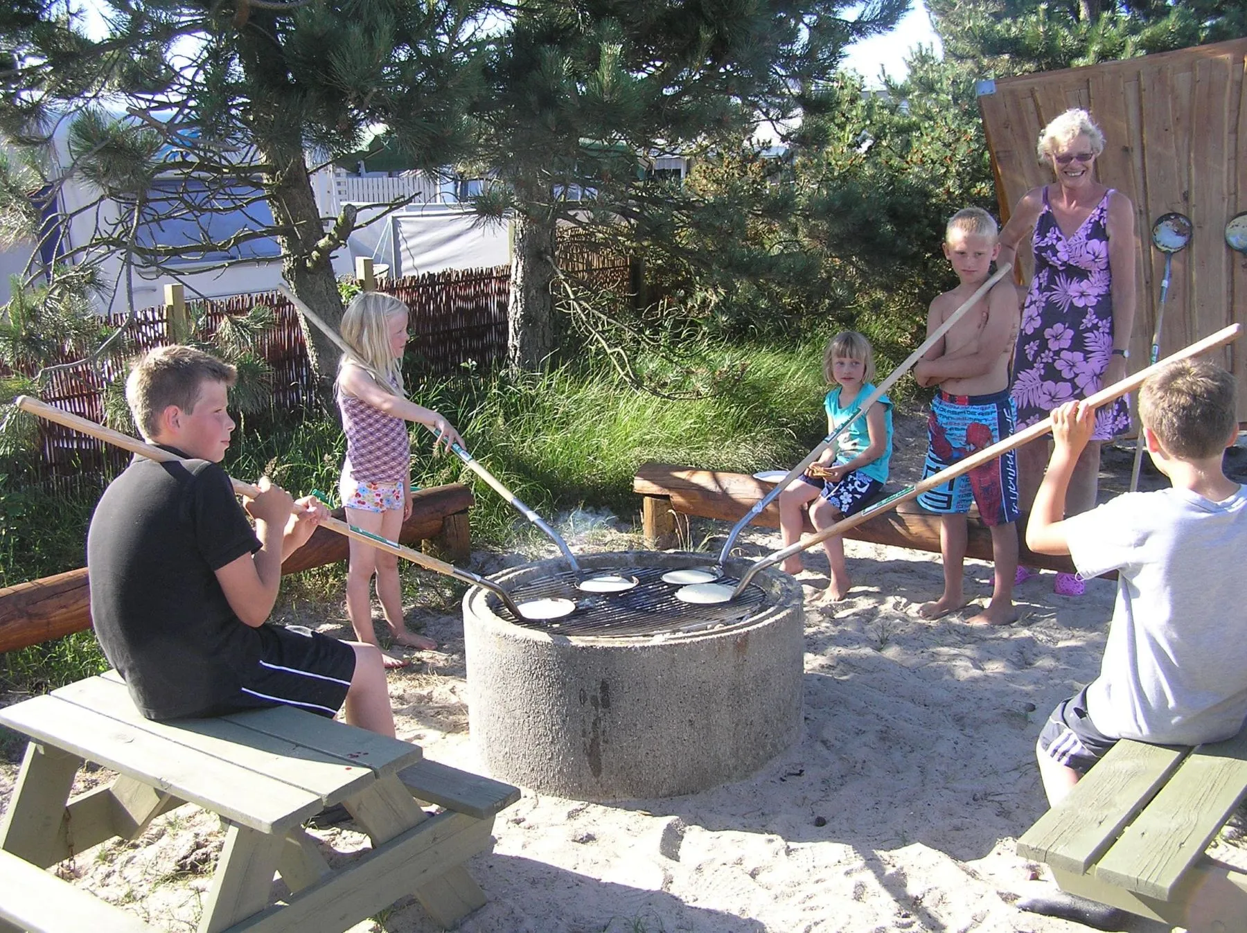 Kinder und eine Frau backen Pfannkuchen über einem Feuer im Freien mit Pfannkuchenpfannen an langen Stielen.