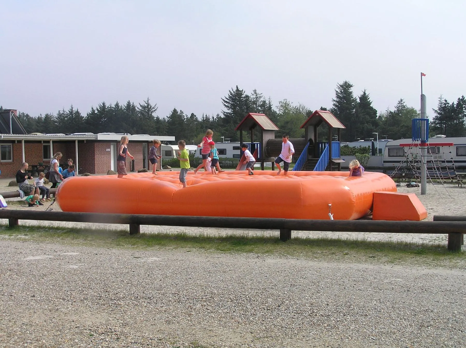 Kinder spielen und springen auf einem großen orangefarbenen aufblasbaren Hüpfkissen auf einem Spielplatz.