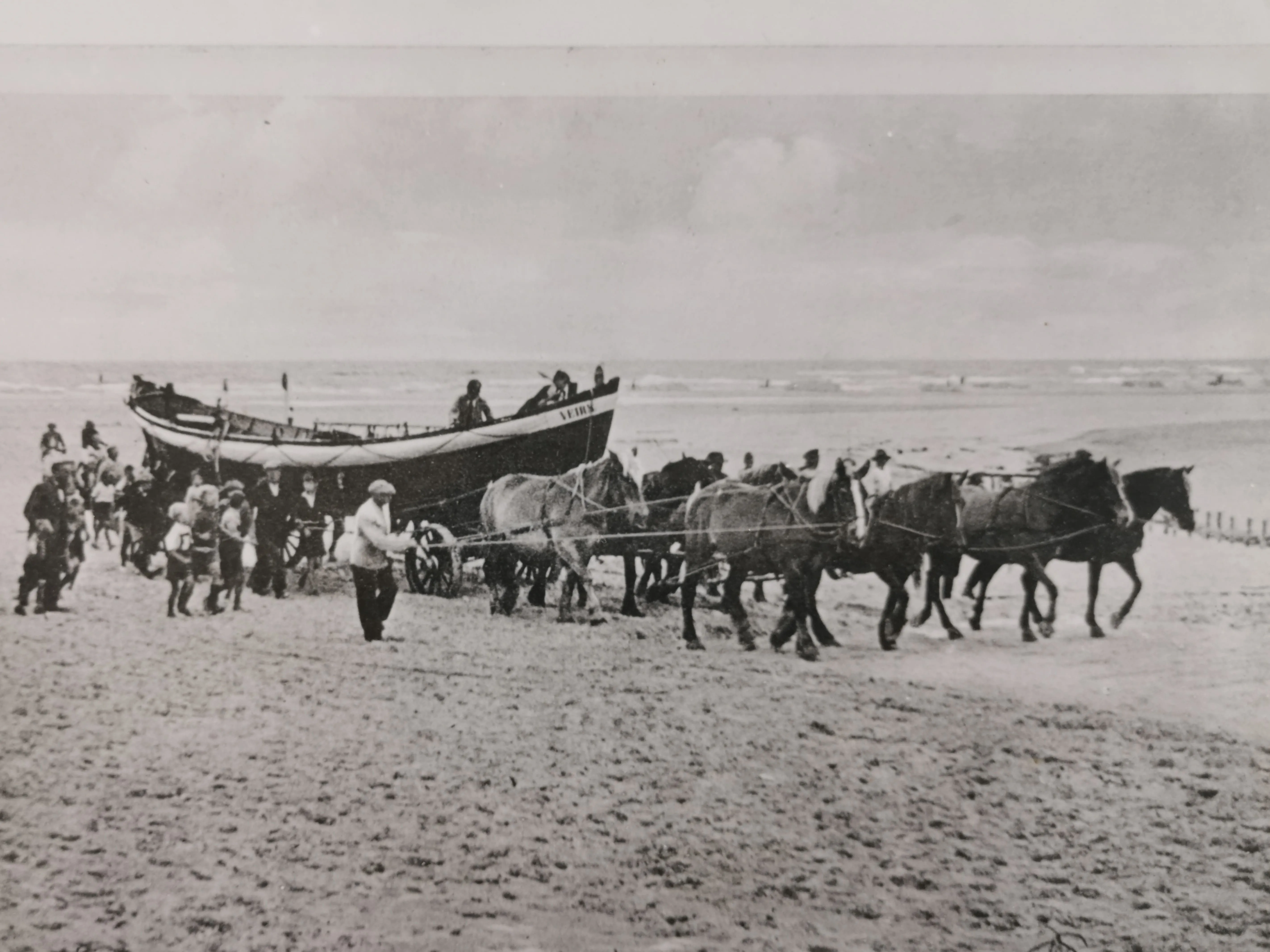 Schwarz-Weiß-Foto aus der Geschichte, das eine große Gruppe von Menschen, Pferde und ein Boot am Strand zeigt.