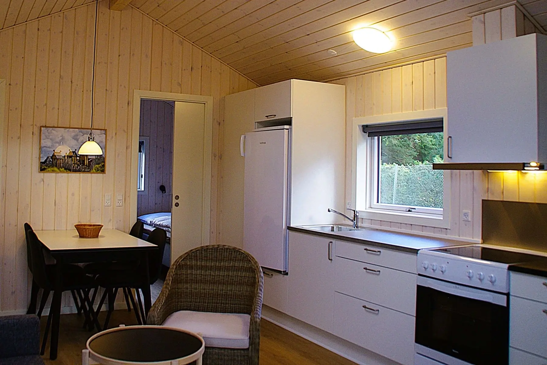 Lyst køkken med hvide skabe, køleskab, komfur og vindue med udsigt til grønne buske, samt en spisestue med bord, stole og en dør til et soveværelse.