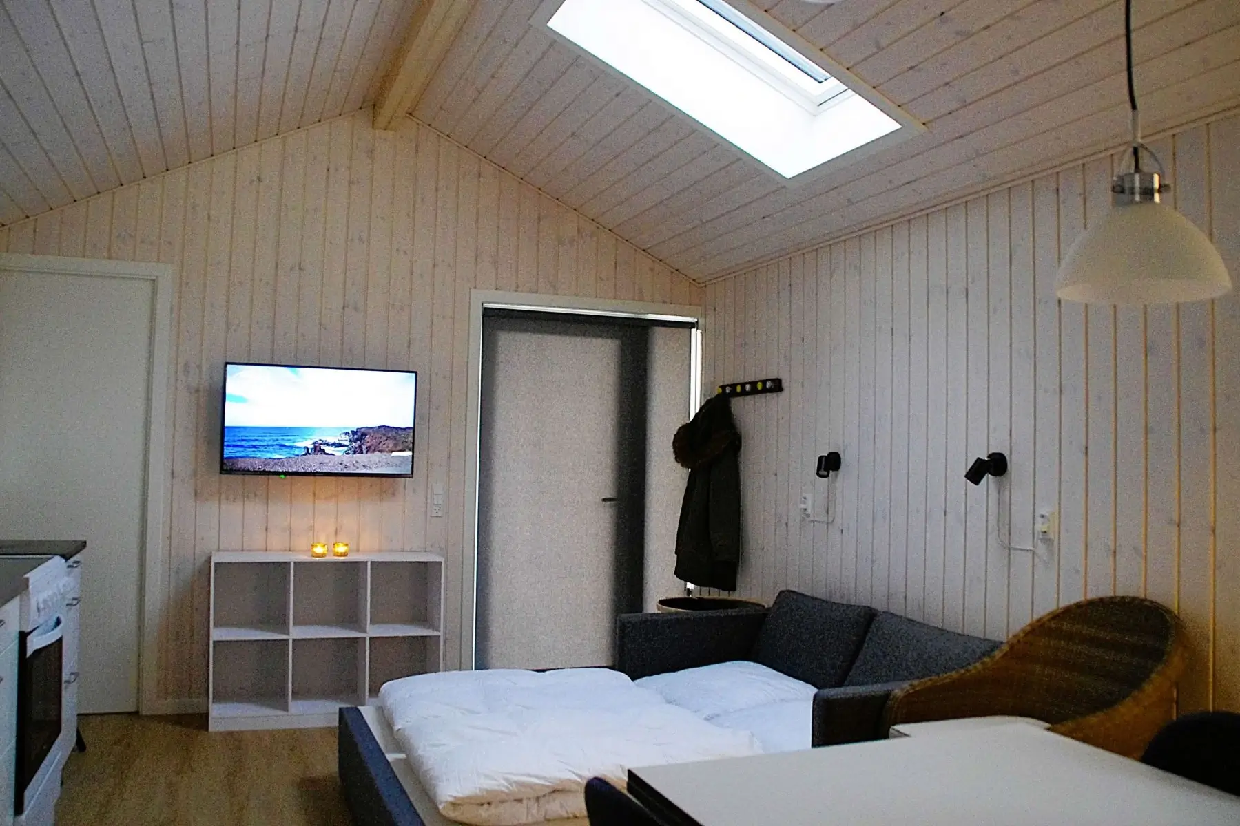 Lyst værelse med snekkehulsscene på væg-tv, foldesofa med sengetøj, hvidt reolsystem, loftvindue og væghængt lampe.