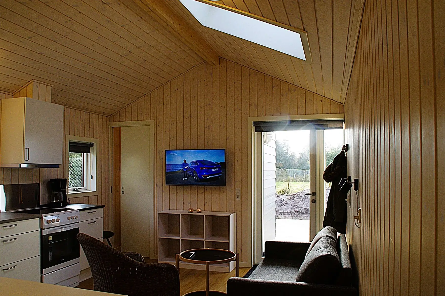 Lyst træbeklædt rum med køkkenområde, fladskærms-tv, grå sofa og glasdør til udendørsområde.