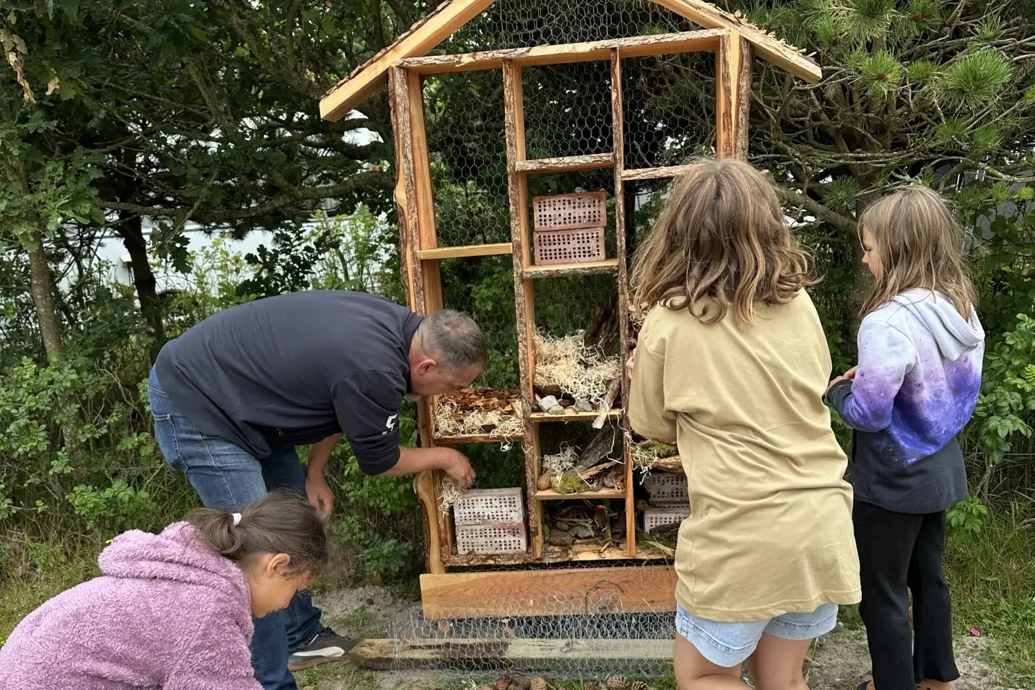 En mand og tre børn interagerer med et insektbo lavet af træ og naturmaterialer i en udendørs have.