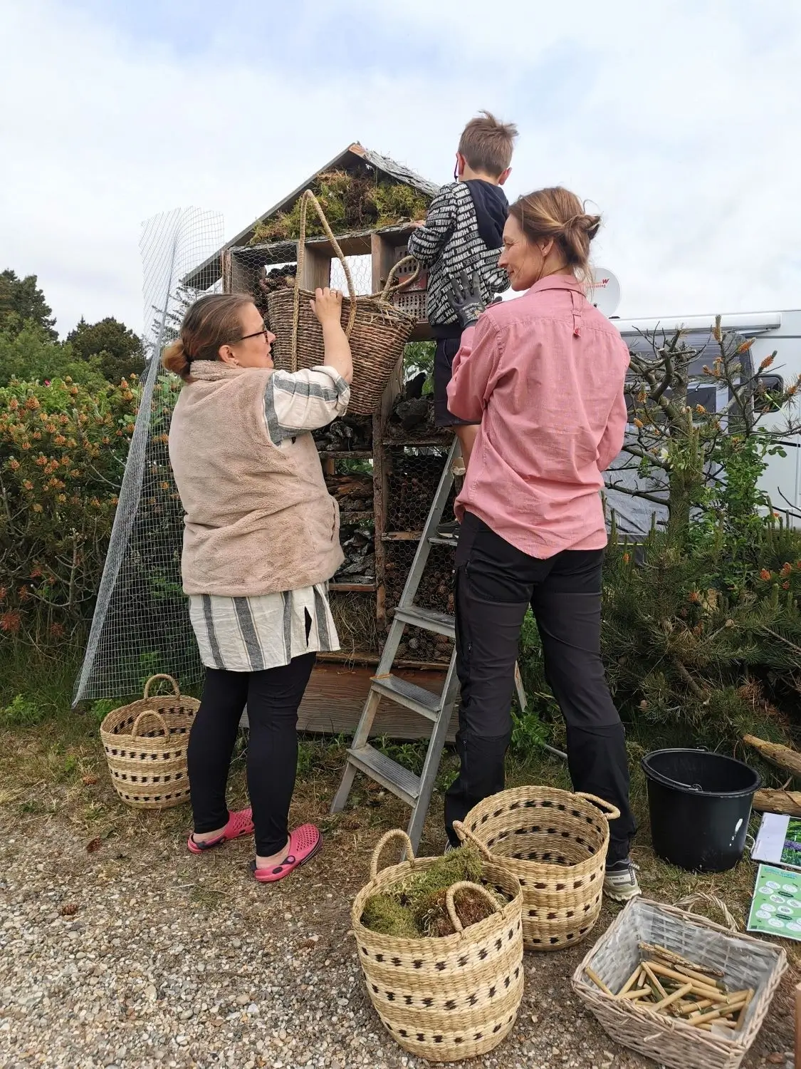 To kvinder og et barn ved et insekthotel, hvor de fylder kurve med naturmaterialer på en grusvej omgivet af buske.