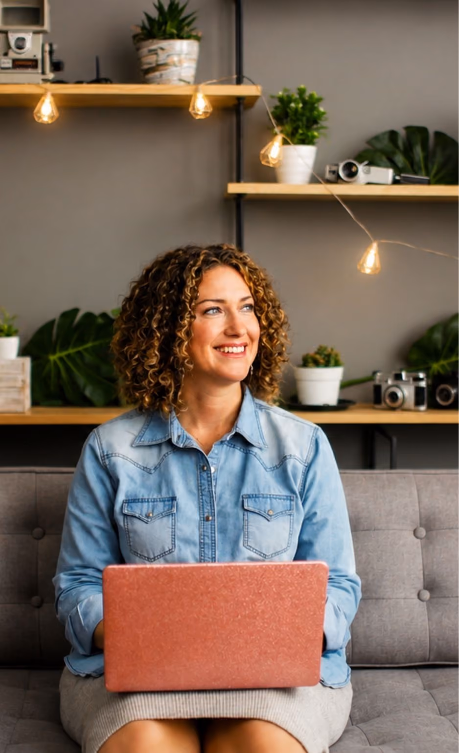 Smiling woman with curly hair in denim shirt sitting on couch using a laptop, with plants and cameras behind her.