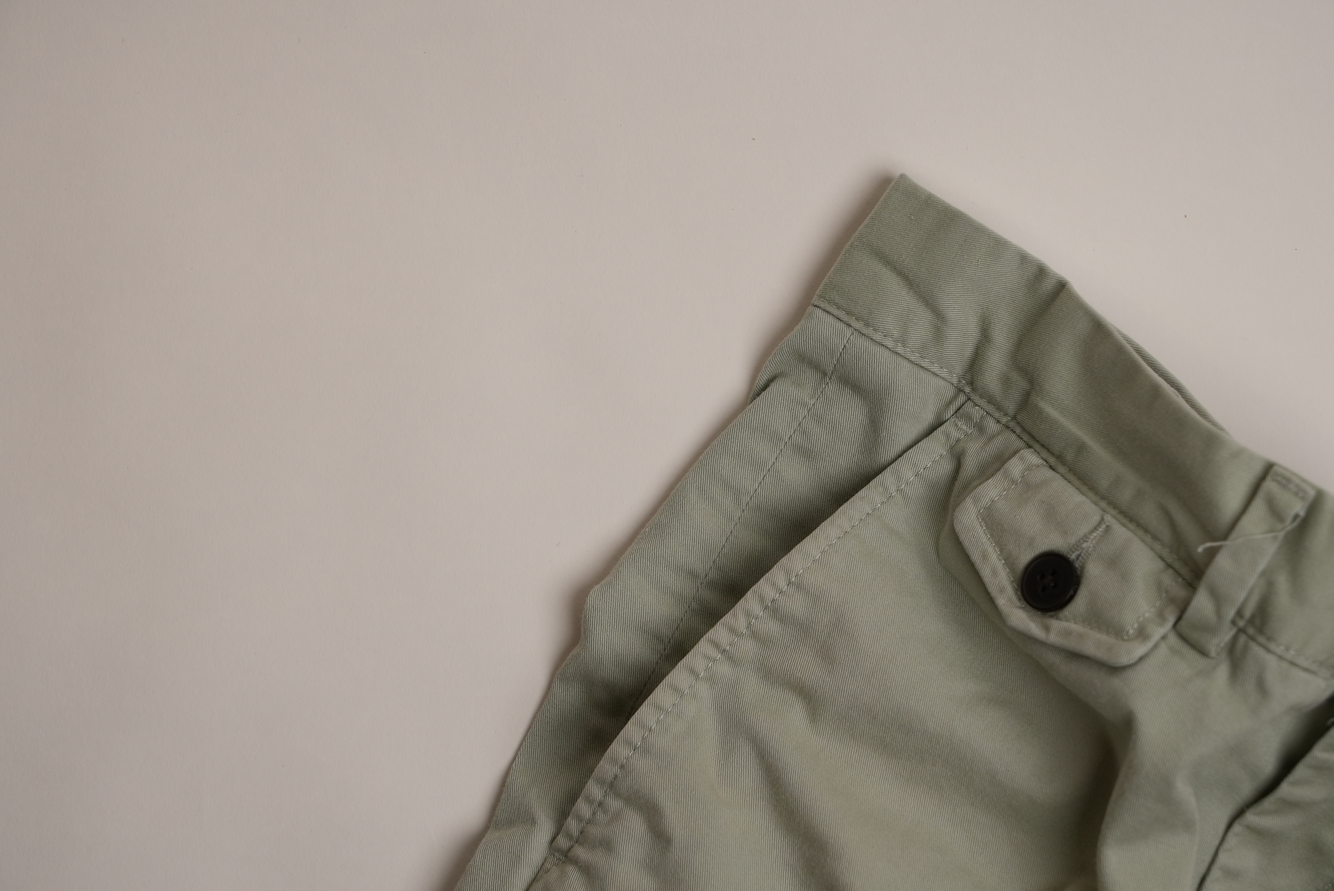 Close-up of the waistband and pocket area of light green pants with a black button on a beige background.