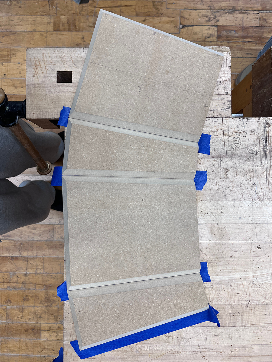 Four rectangular wooden boards with beveled edges taped together with blue painter's tape on a wooden workbench.