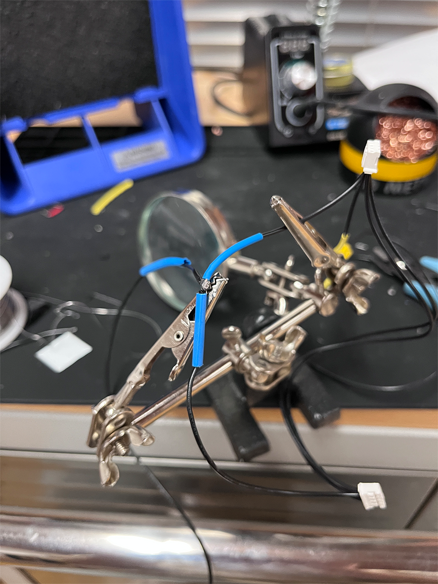Helping hands tool holding black and blue wires with soldered connections on a workbench.