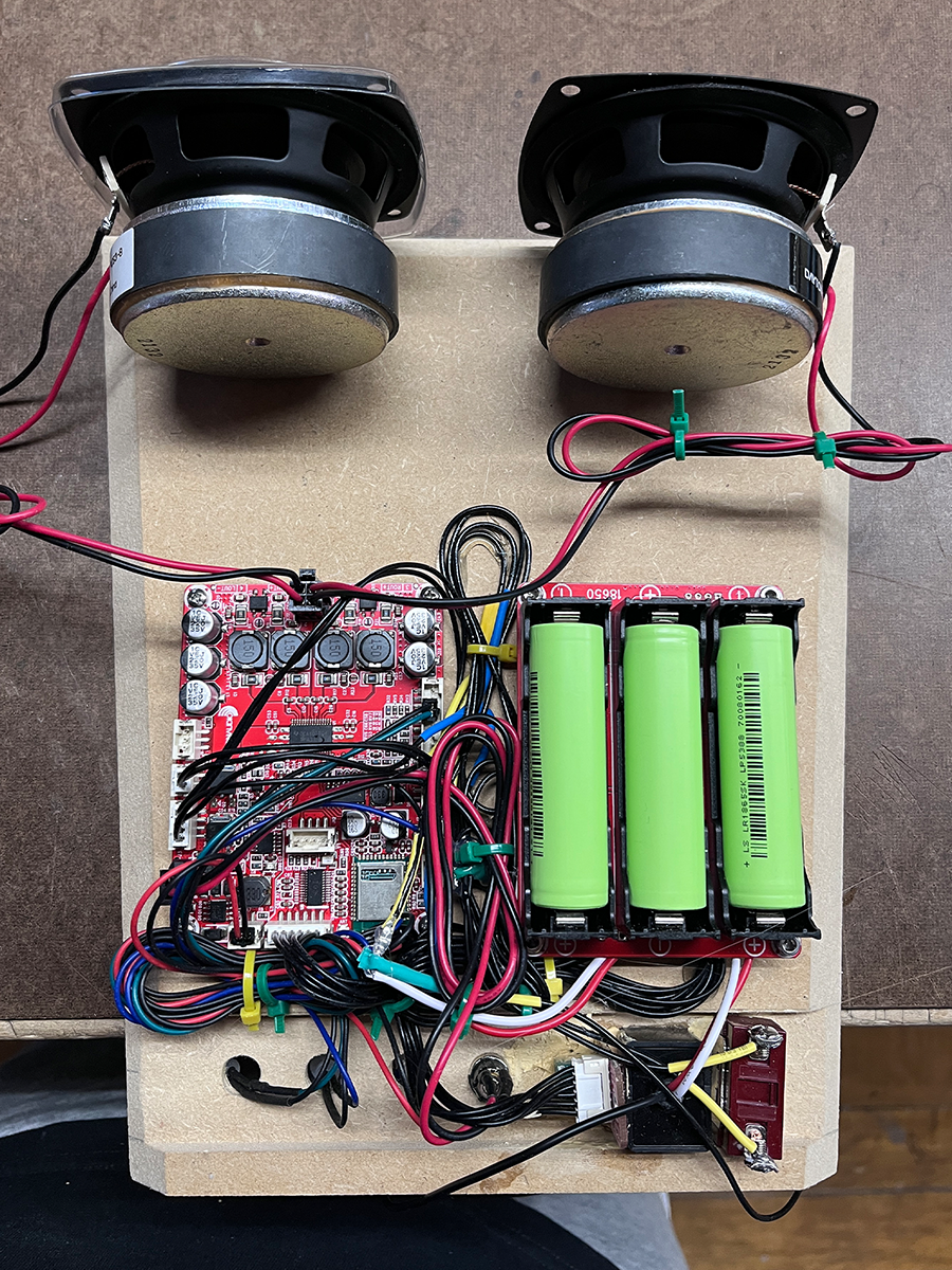 Two speakers, a circuit board with capacitors, and three green cylindrical batteries connected with red, black, and other colored wires mounted on a wooden board.