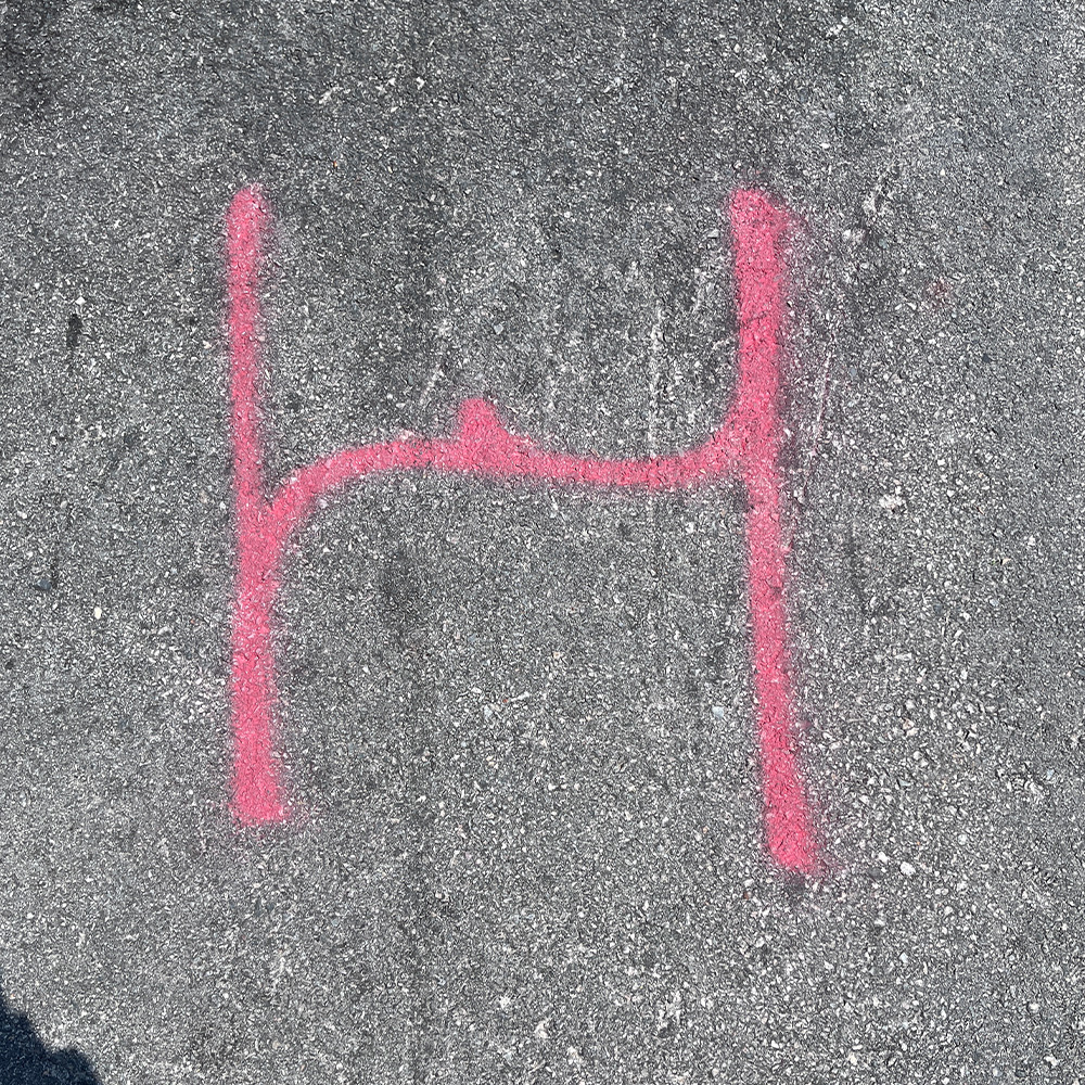 Pink spray-painted symbol resembling the letter 'H' on gray asphalt pavement.