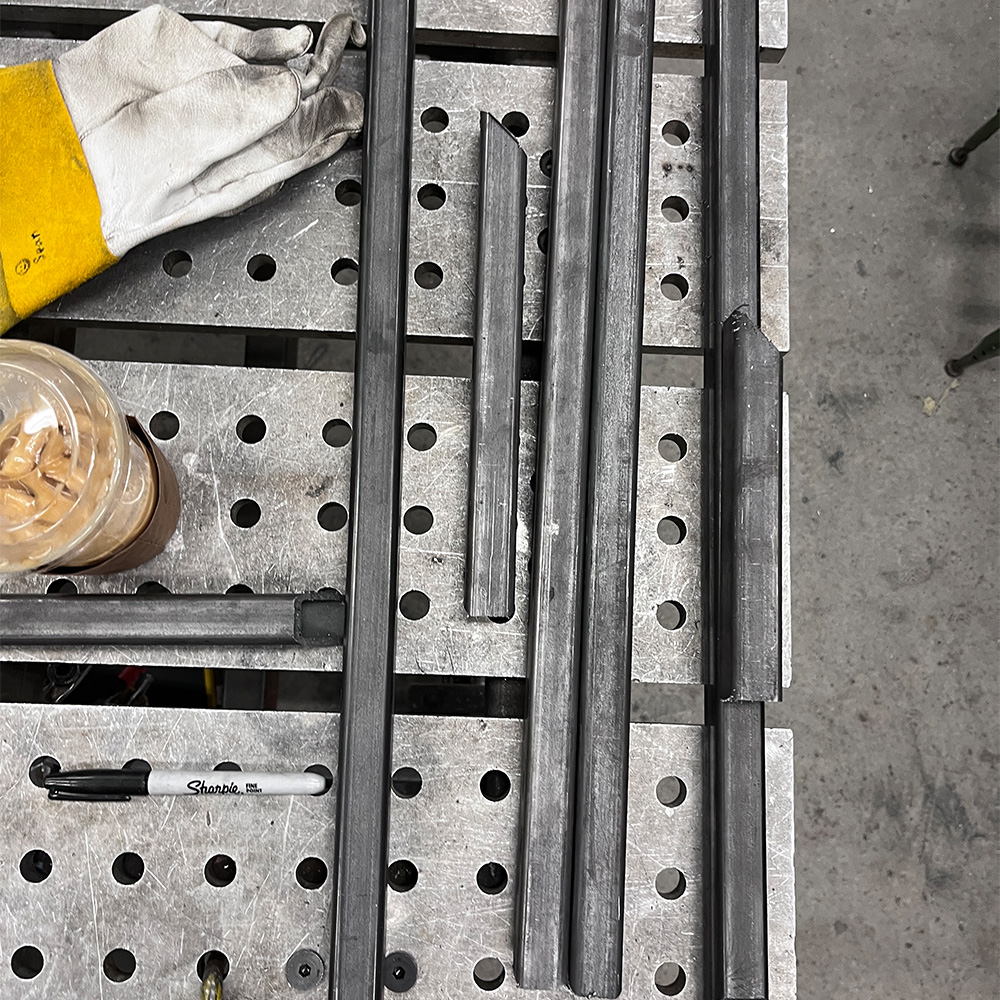 Steel rectangular tubes cut at angles arranged on a perforated metal workbench with a work glove, iced coffee, and black Sharpie marker.