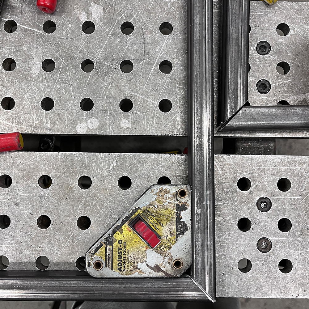Corner of metal square tubing clamped on a perforated metal workbench with a worn magnetic welding square tool.