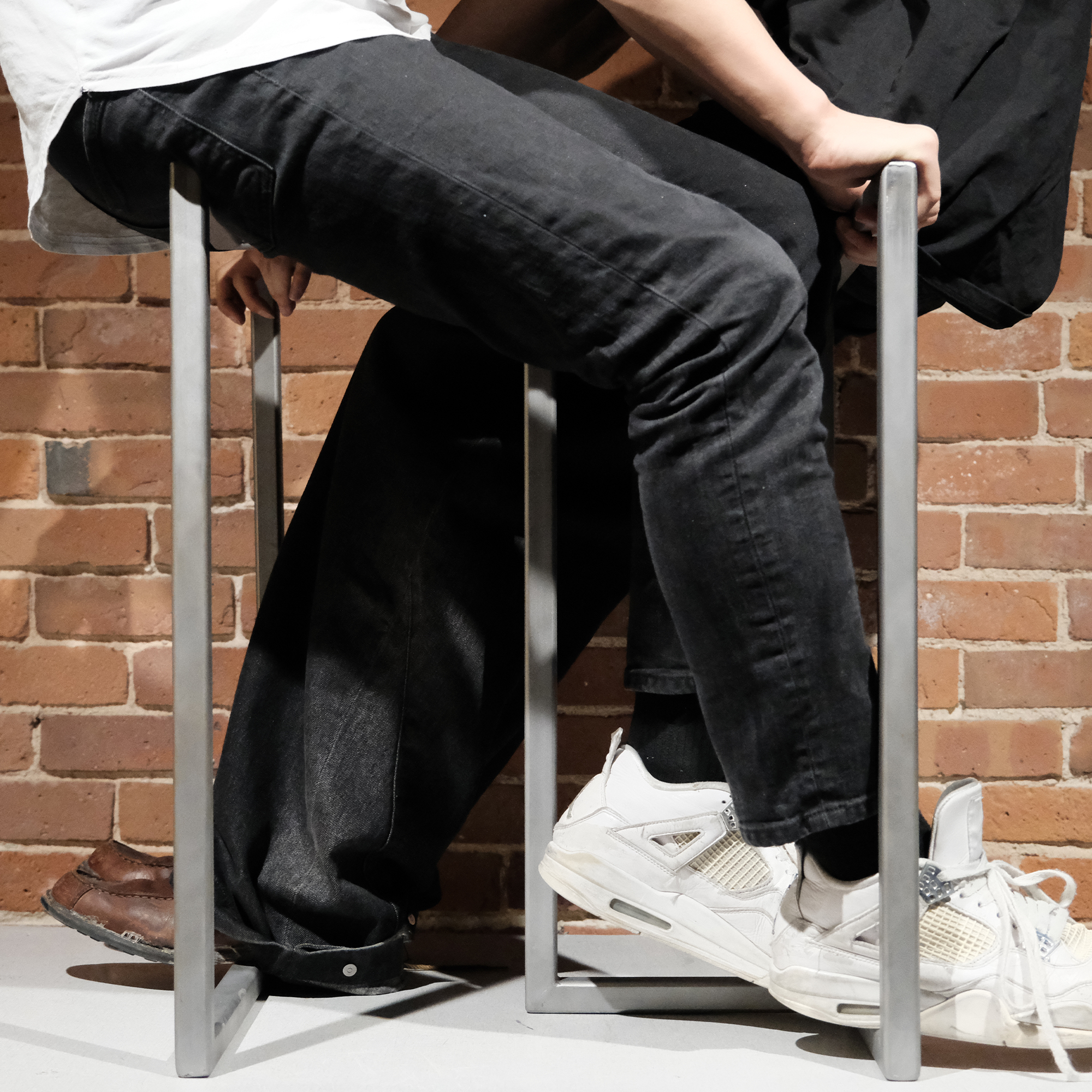 Lower legs and feet of two seated people against a brick wall, one wearing white sneakers and the other brown shoes.