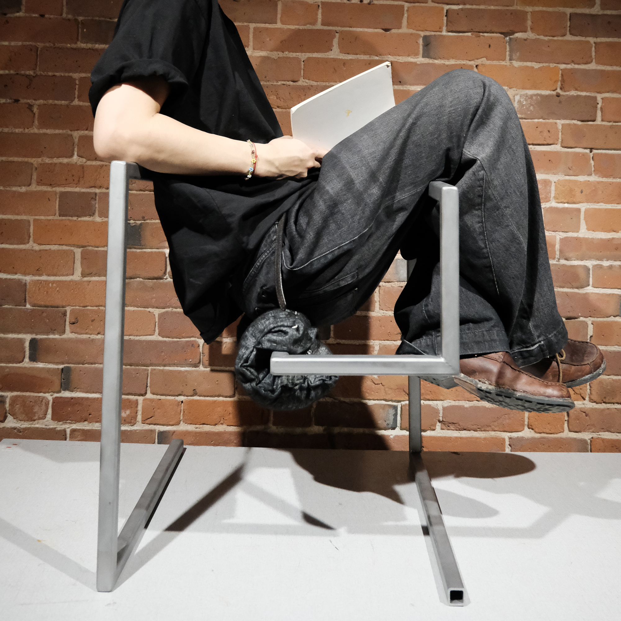 Person in black shirt and jeans balancing on a modern metal chair against a brick wall.