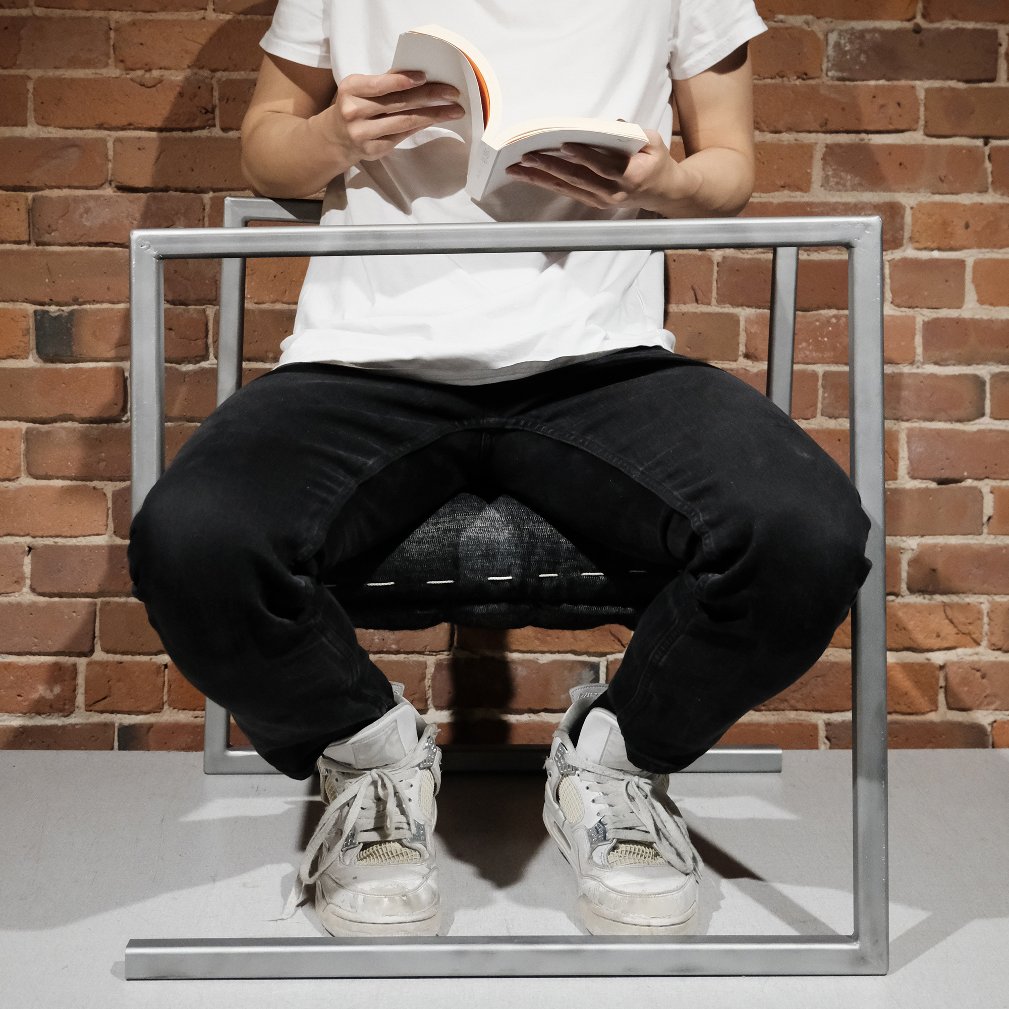 Person wearing black pants and white sneakers sitting on a chair reading a book in front of a brick wall.