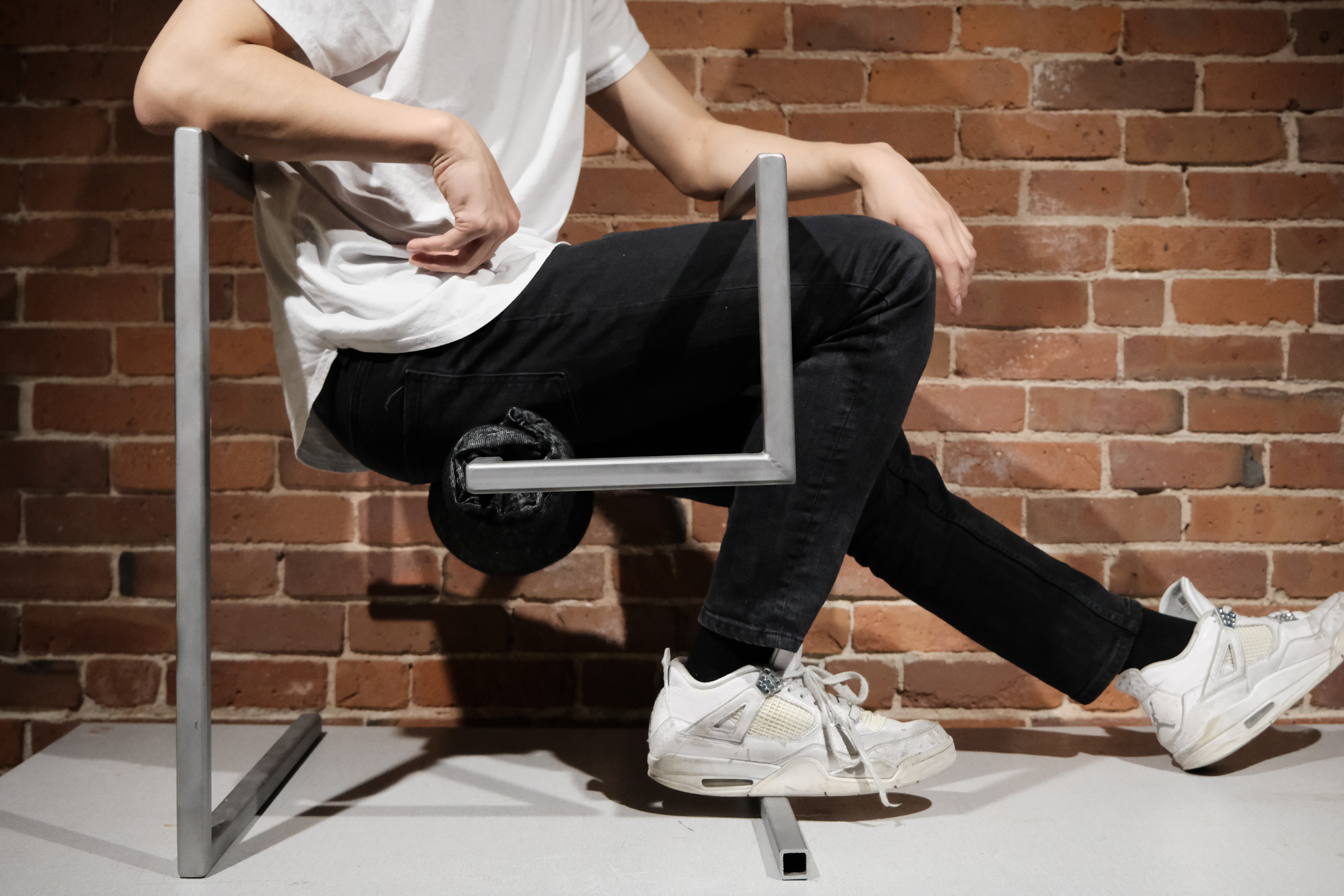 Person sitting on a metal chair with a black cushion under the seat, wearing black pants, white sneakers, and a white shirt against a brick wall.