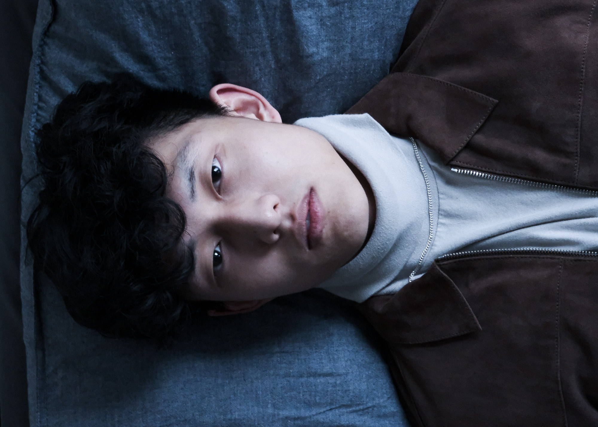 Young man with curly hair lying on a blue surface, wearing a light turtleneck and brown jacket.
