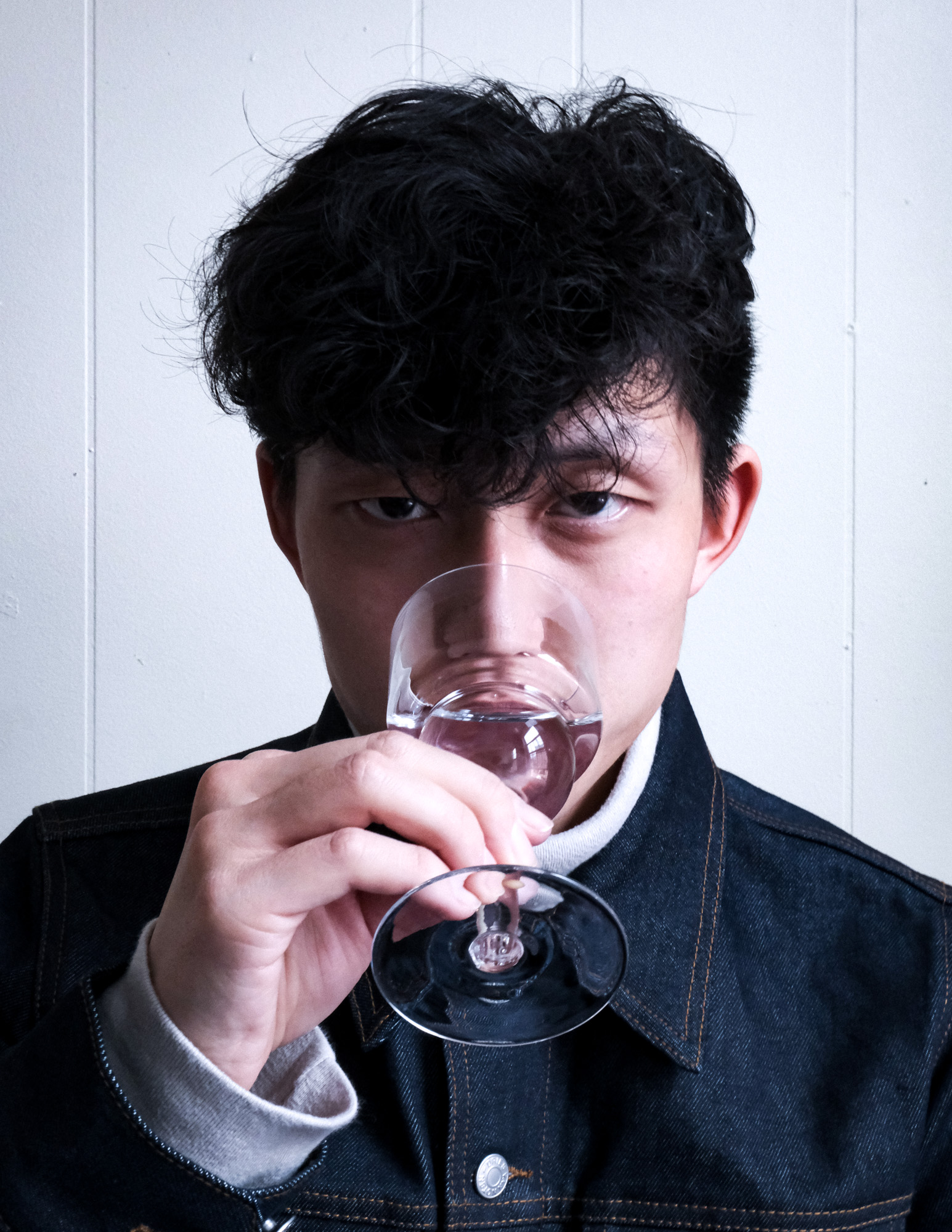 Person with dark curly hair wearing a dark jacket, holding and sipping from a clear wine glass.