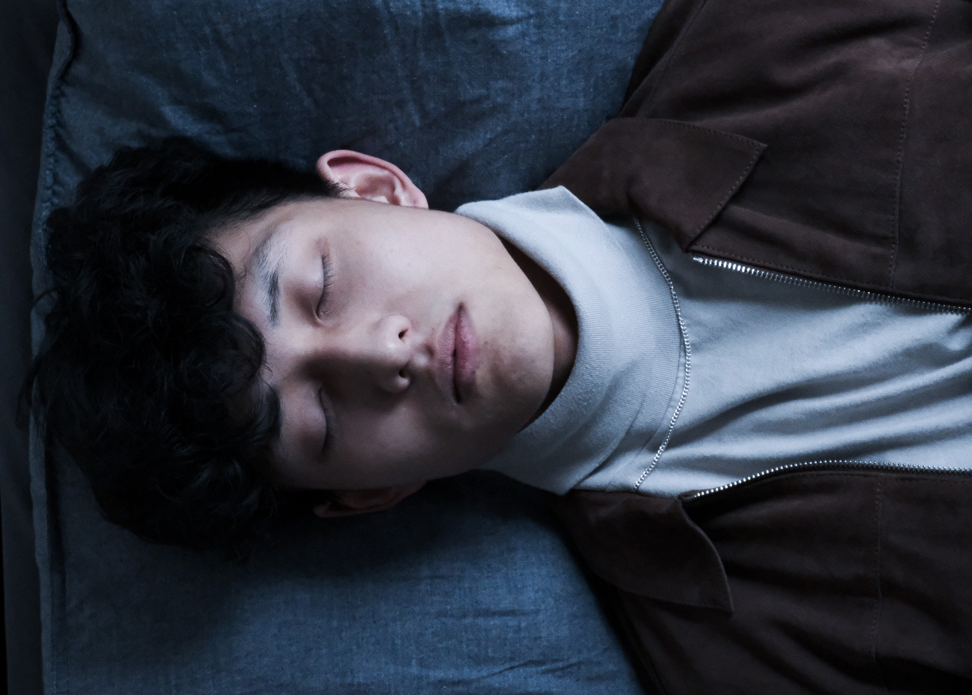 Young man with curly hair lying on a blue surface with eyes closed, wearing a white turtleneck and brown jacket.